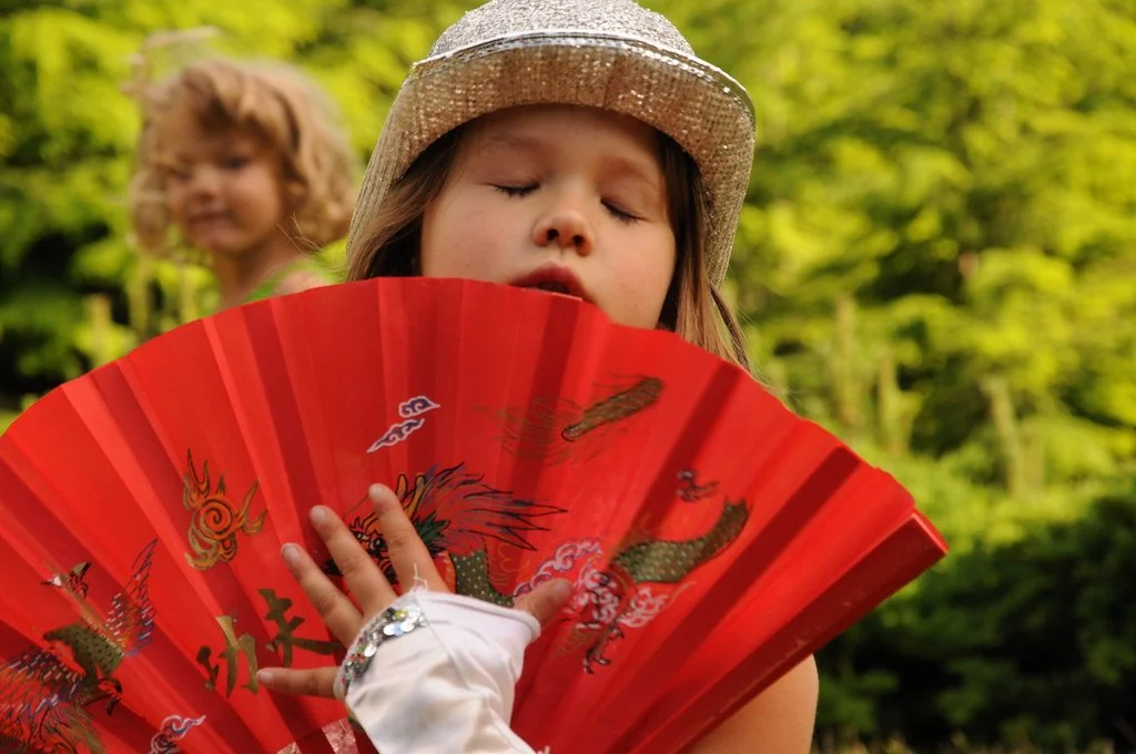 A young girl holding a red decorative fan with a dragon design, wearing a straw hat, with her eyes closed, in a blurred outdoor setting with green trees in the background. A child with curly hair is in the background.