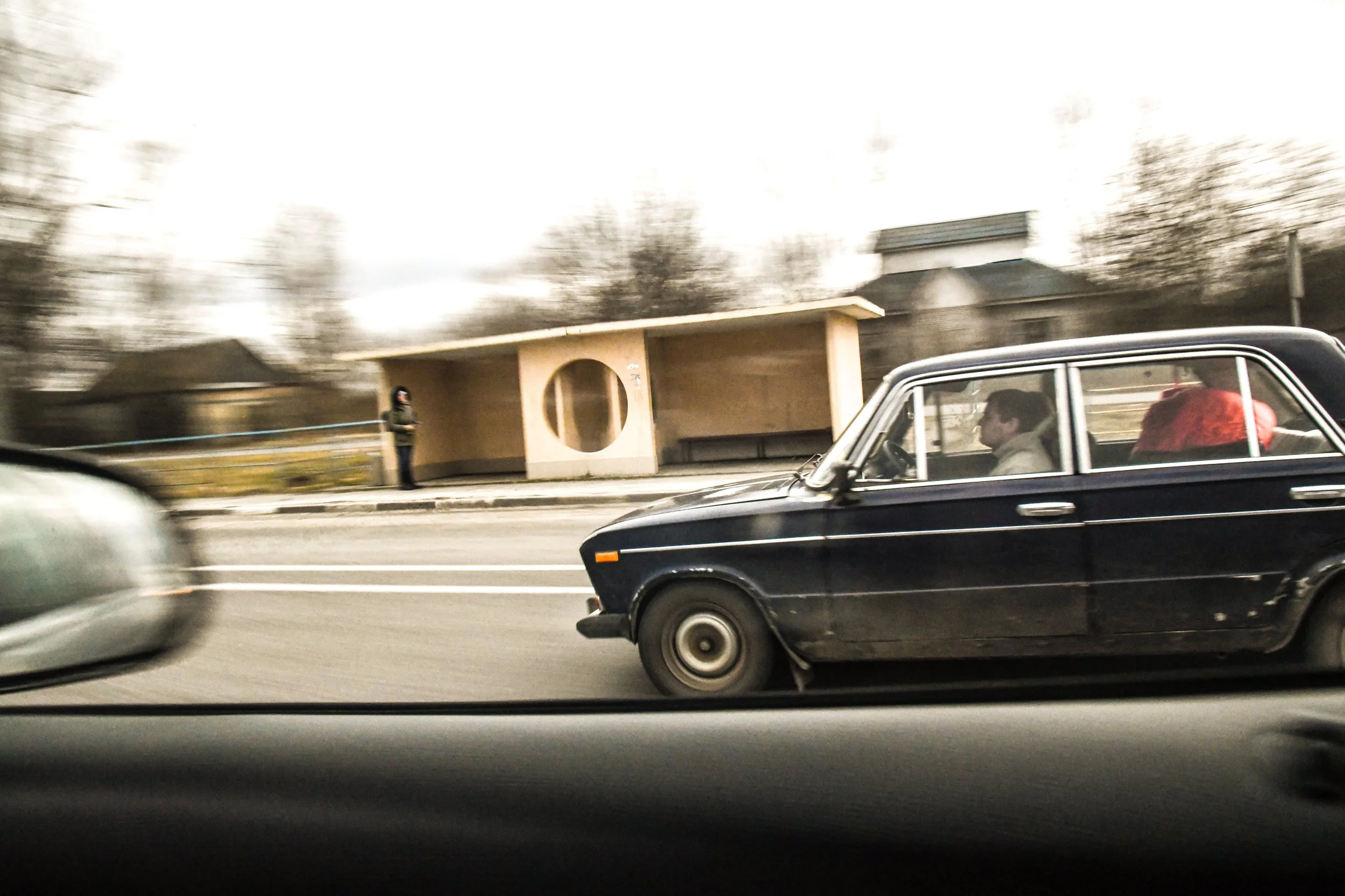 A blue vintage car driving on a road with a person sitting inside and a person holding a red object in the back seat, while a woman stands at a bus stop in the background on a cloudy day.