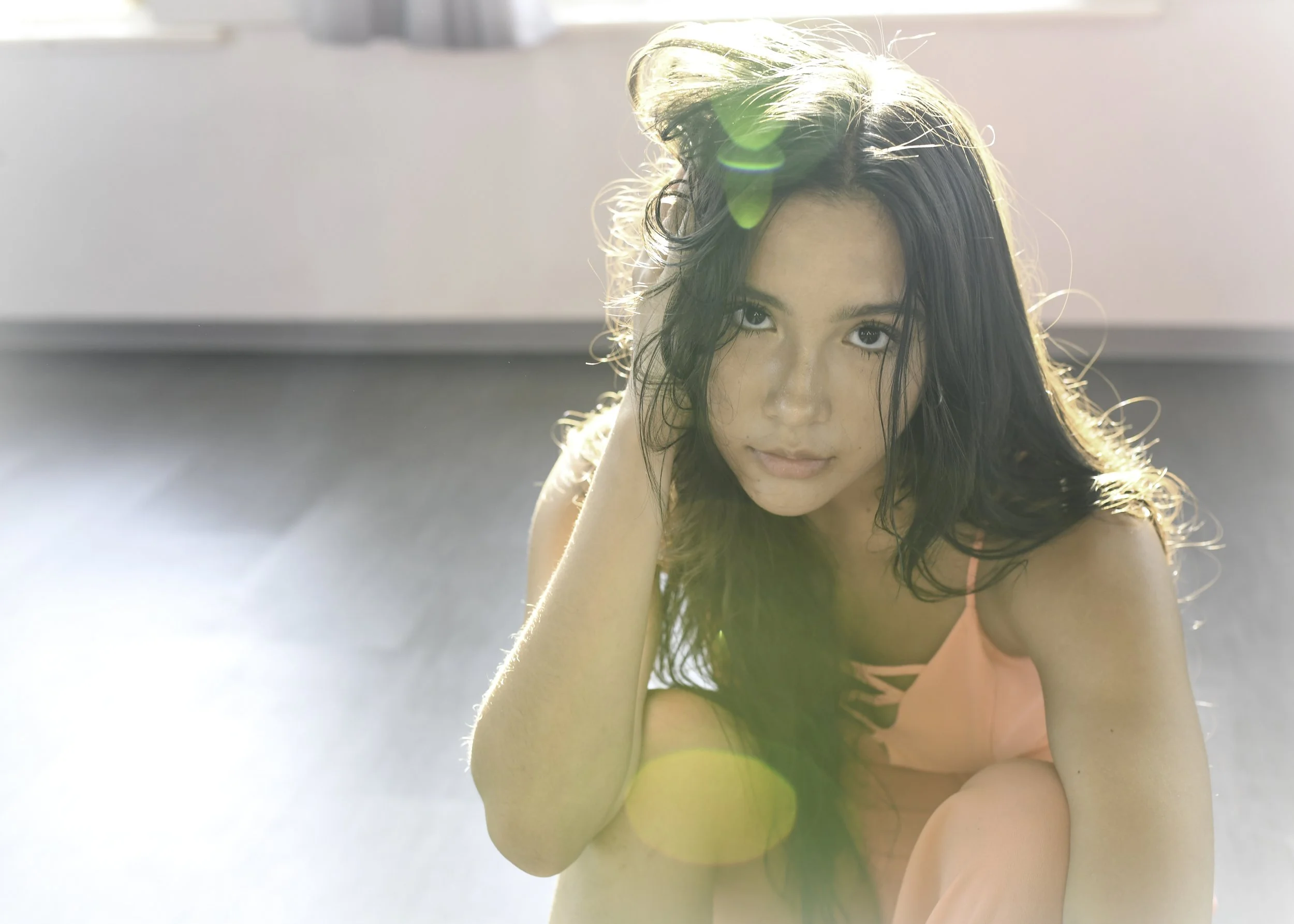 A young woman with dark, wavy hair and blue eyes, wearing a peach-colored top, sitting on the floor with sunlight creating lens flare and backlighting.