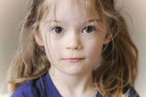 Close-up of a young girl with long, curly blonde hair and blue eyes, wearing a blue shirt.