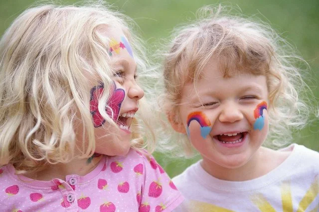 Two young girls with face paint of rainbows and clouds laughing outdoors.