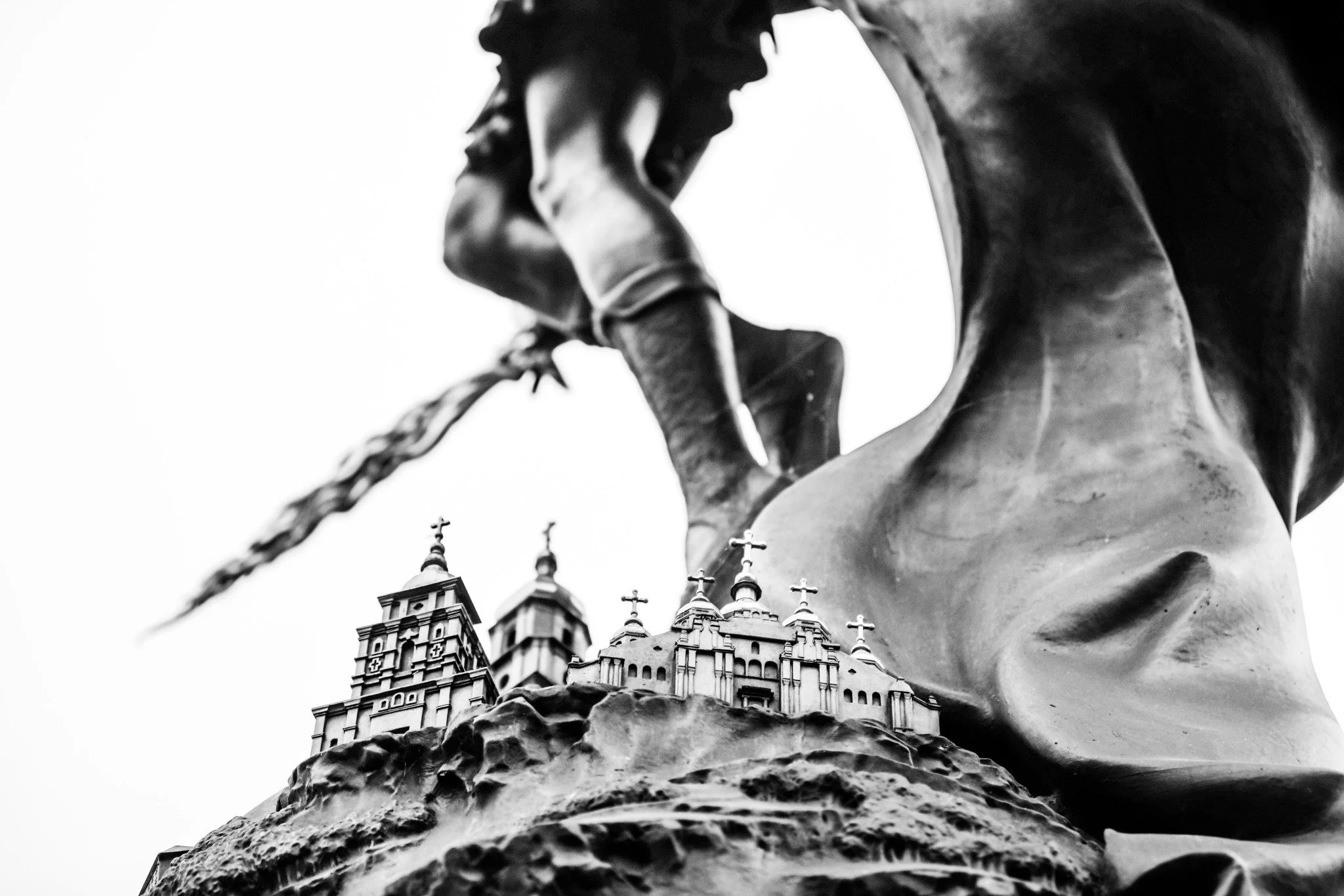 Black and white close-up photograph of a religious statue with a church with multiple crosses on top of its domes in the background under an overcast sky.