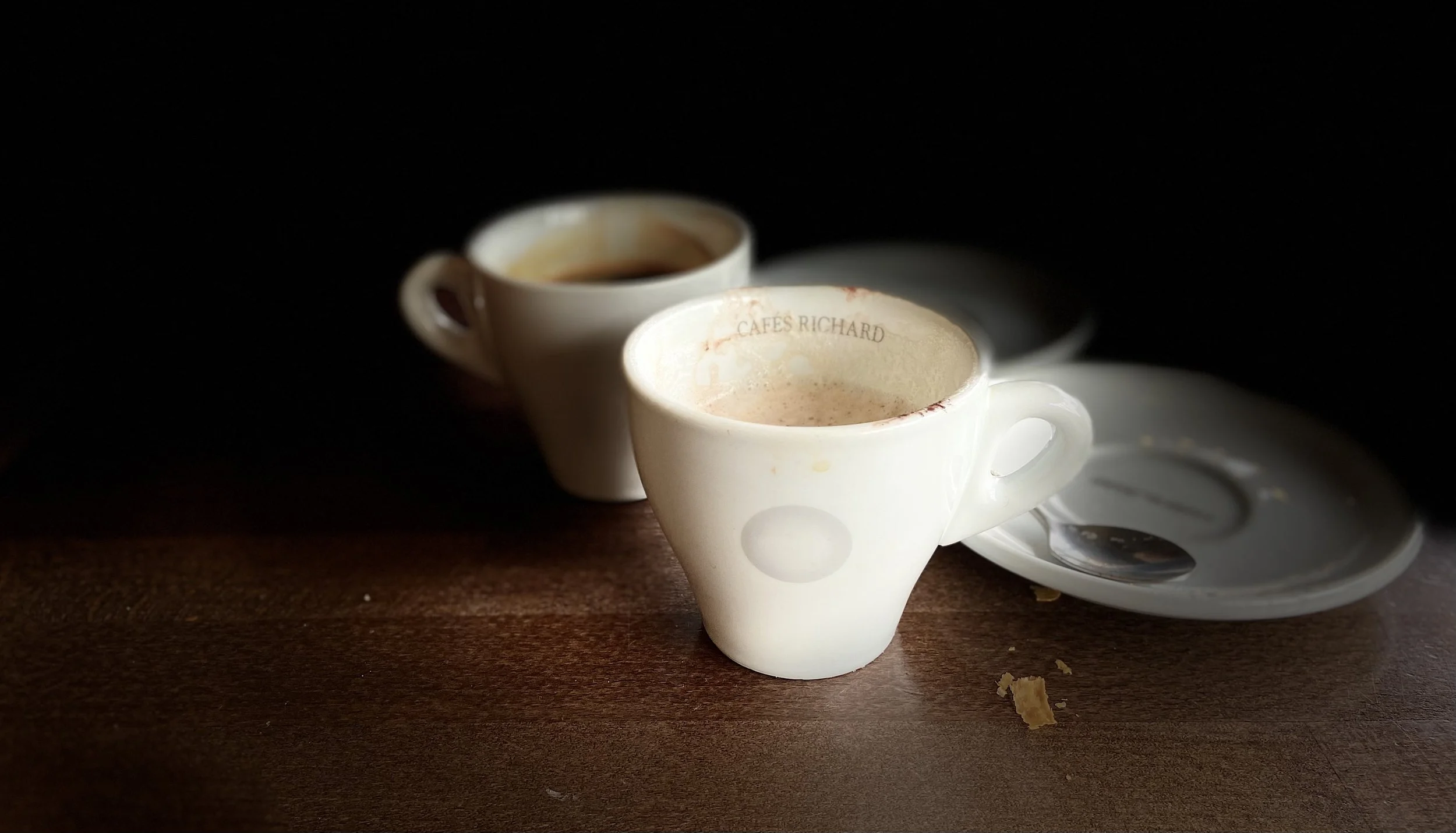 Two white coffee cups with coffee on a dark wooden table, with a white saucer and spoon nearby, against a dark background.