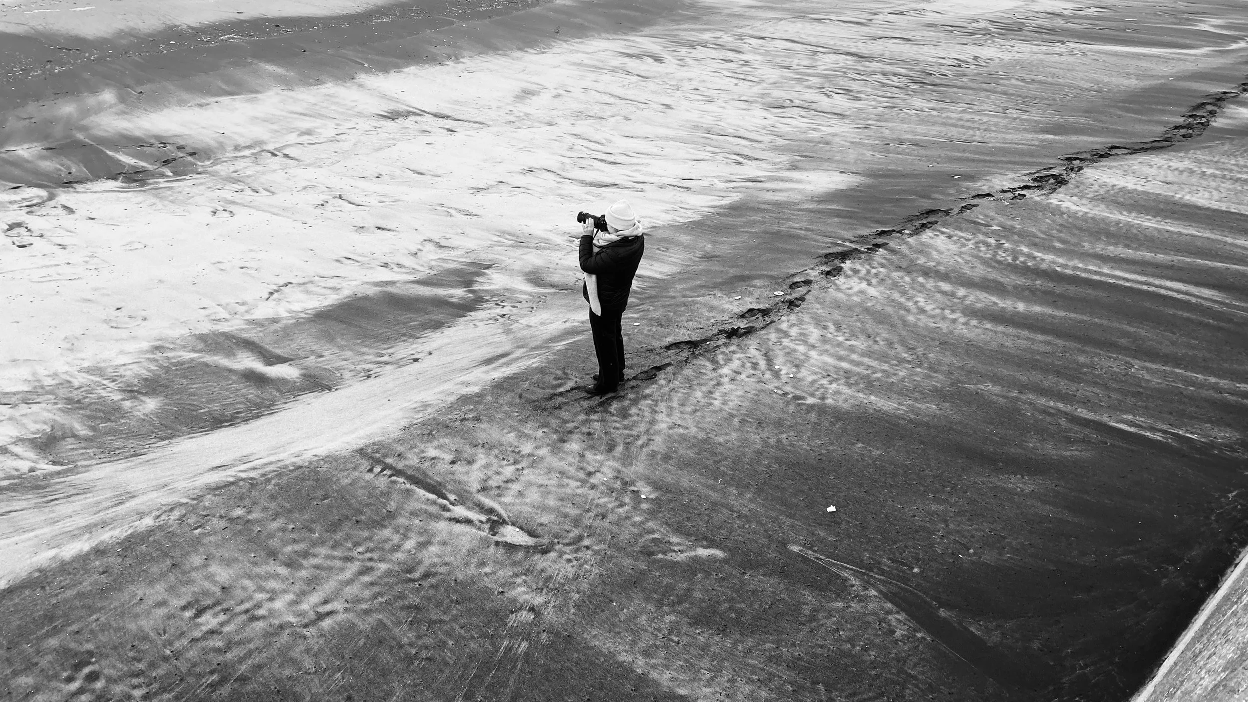 Person standing on snowy beach taking photo of the ocean with a camera, dressed in winter clothing.