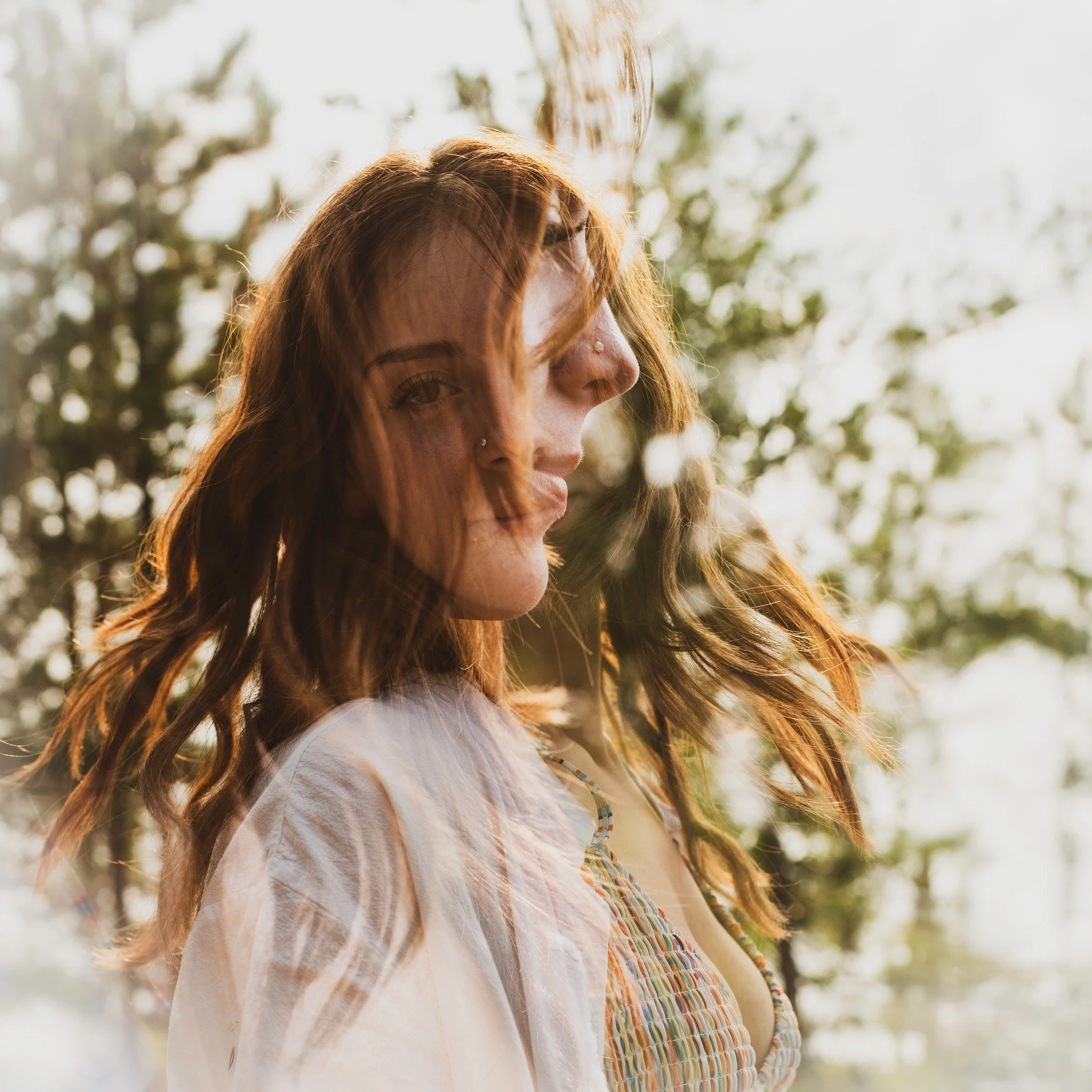 A portrait of a woman with red hair and a nose piercing, looking to the side, outdoors with blurred trees in the background, bathed in warm sunlight.