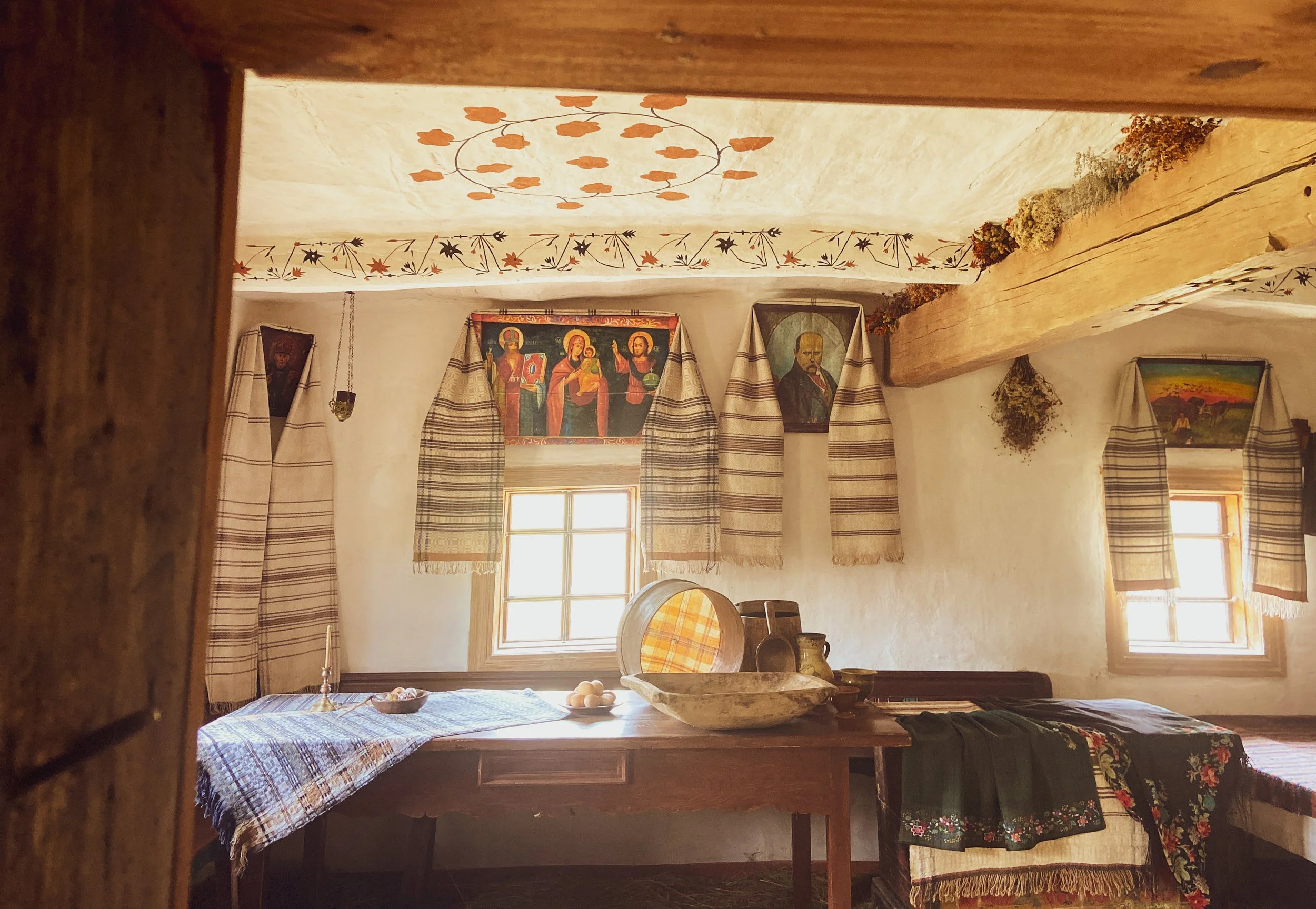 Interior of a traditional rustic room with white plaster walls, wooden beams, and decorative textiles. There are two windows with striped curtains, religious icons and paintings on the walls, and a wooden table covered with woven fabric, bowls, and eggs.