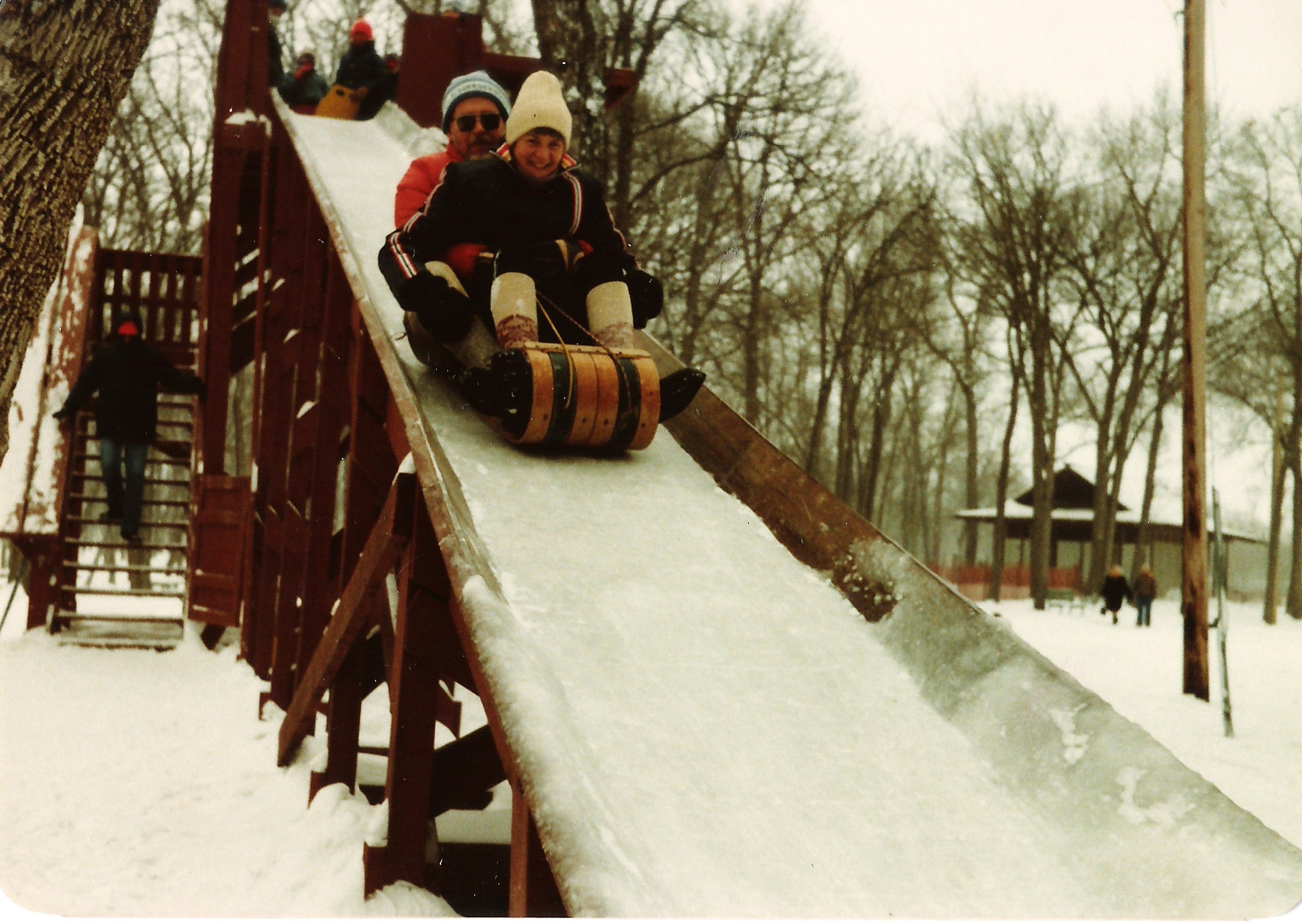 Two children in winter clothing riding a sled down a snowy slide at a playground, with a man in the background at the top of the slide and other children playing nearby.
