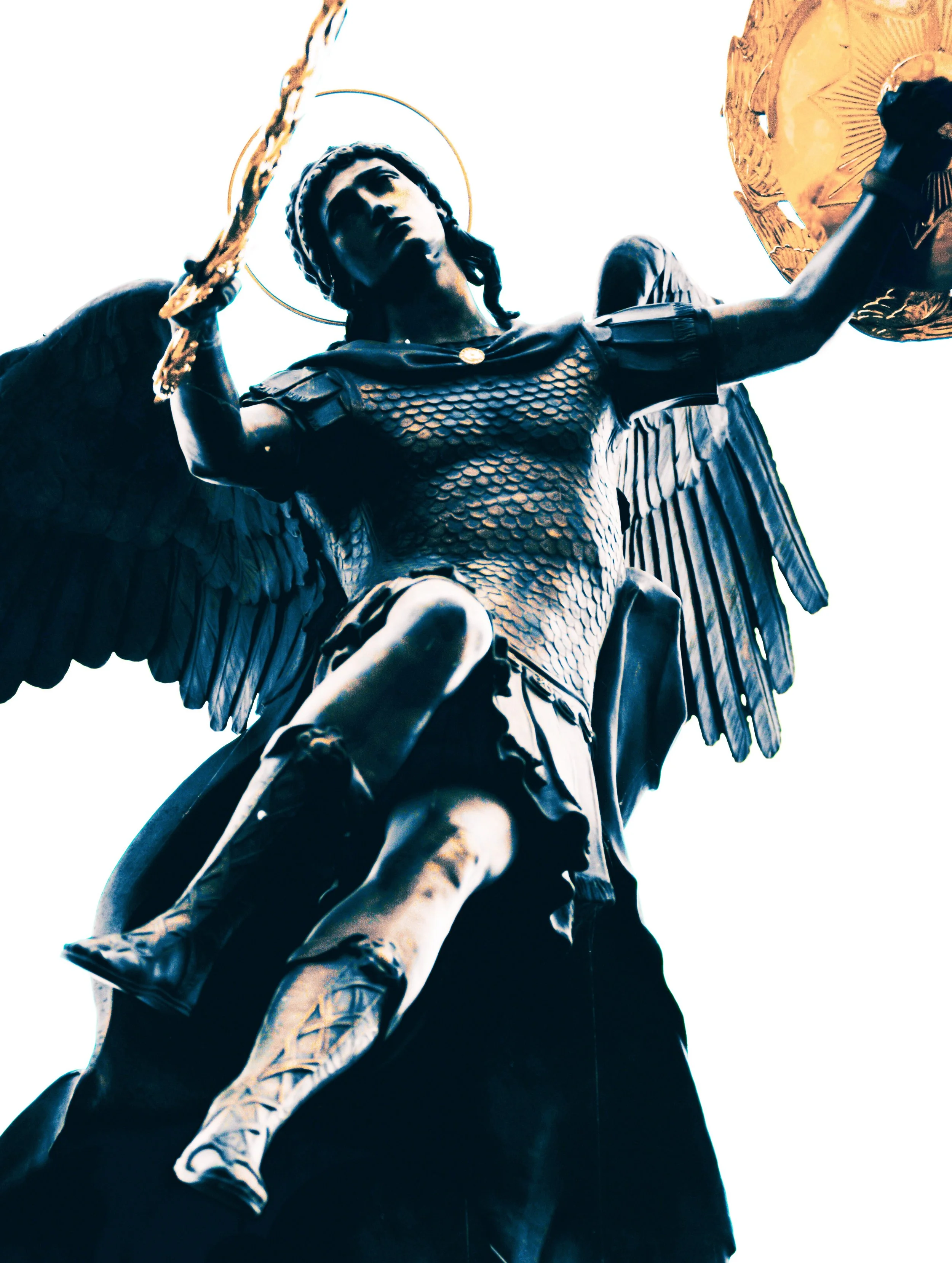 Close-up of a dark-colored statue of a winged figure with a halo, holding a shield and a sword, viewed from below against a bright sky.