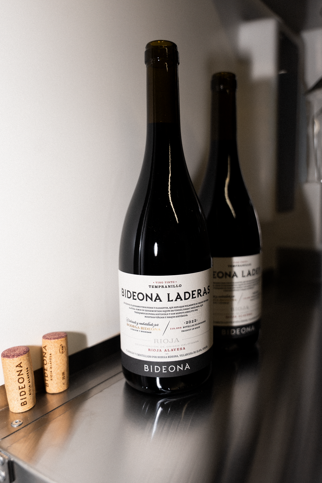 Two bottles of Bideona Laderas red wine on a metal shelf, along with two wine corks, in a storage area.