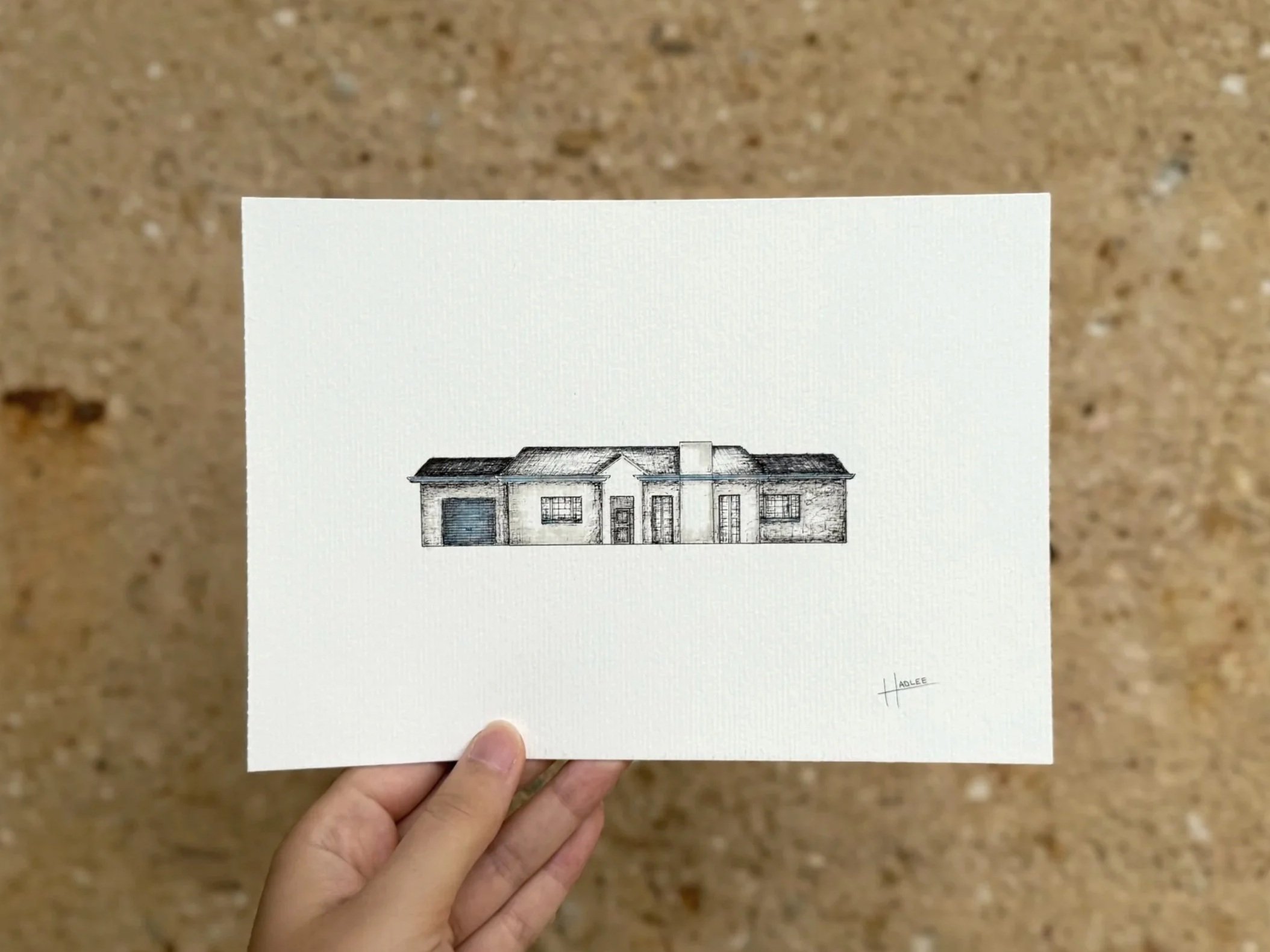 Sketch of a house on a white piece of paper, held by a person over a brown textured surface.