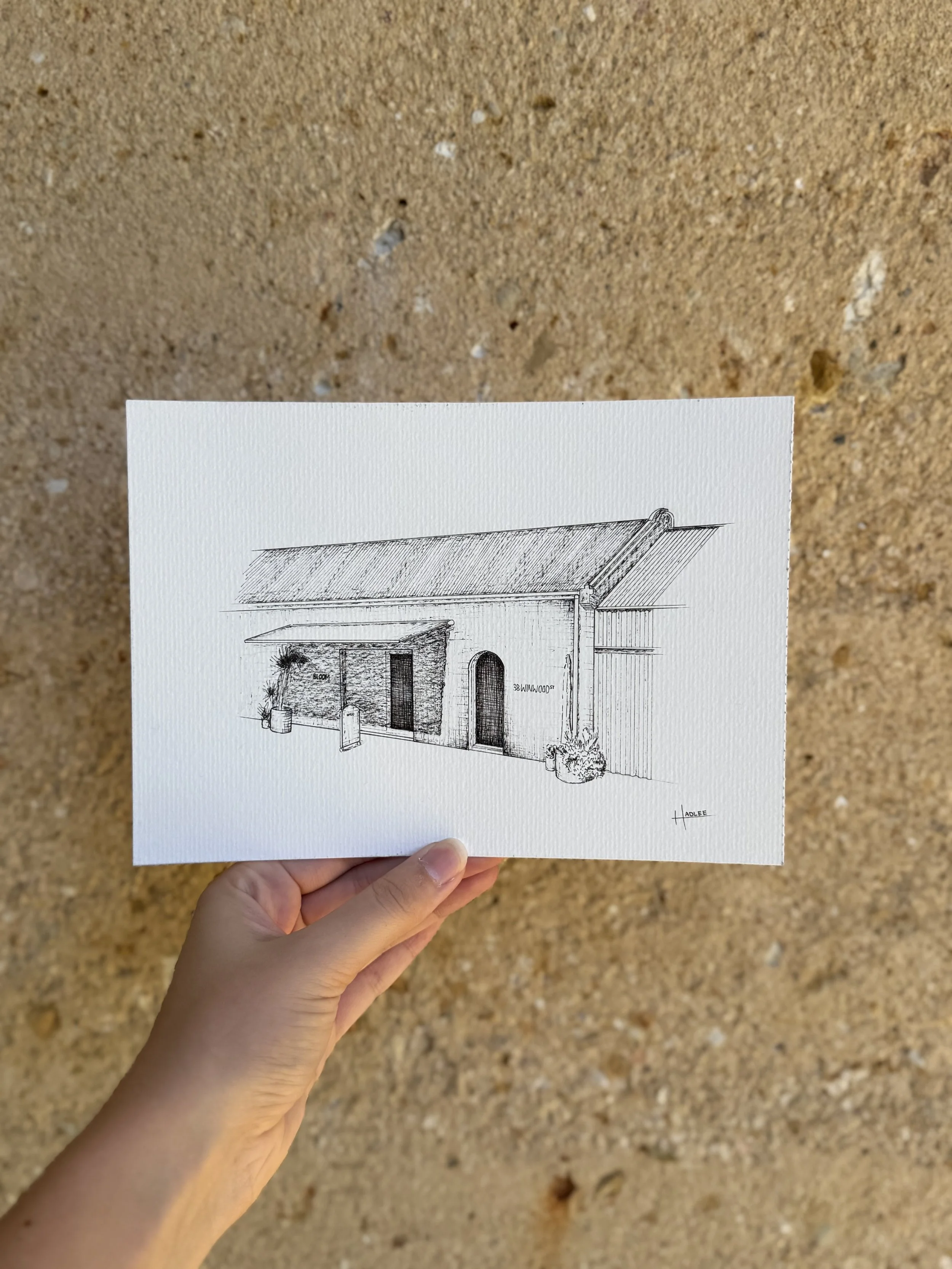 Hand holding a sketch of a building with a sloped roof, arched doorway, and plants outside against a gravel background.