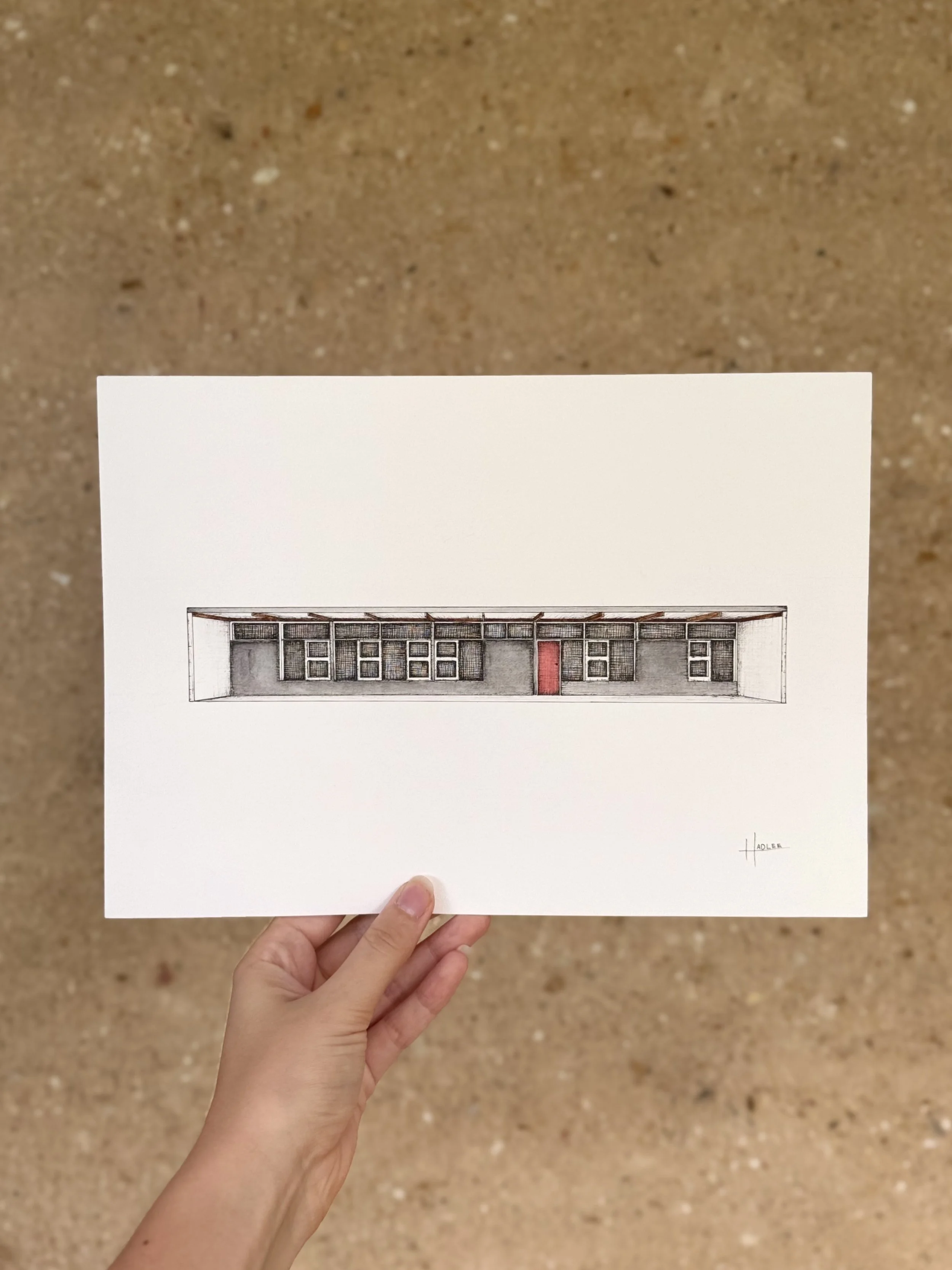 A hand holding a piece of paper with an architectural drawing of a building, featuring a red door, below a roof with exposed beams, set against a brown carpeted floor background.