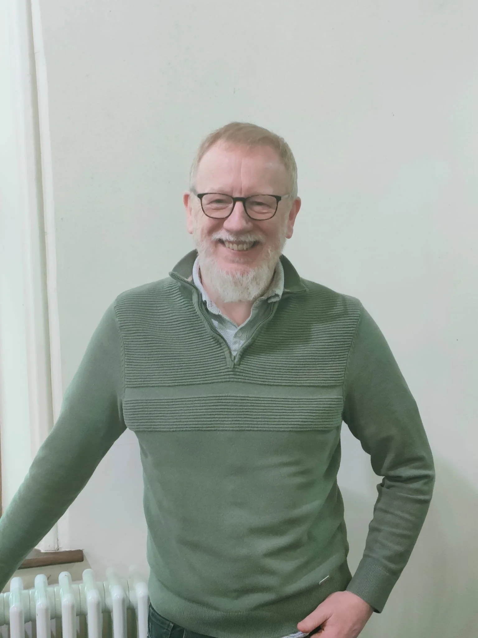 Smiling man with glasses and a beard, wearing a green striped sweater, standing near a white wall and radiator.