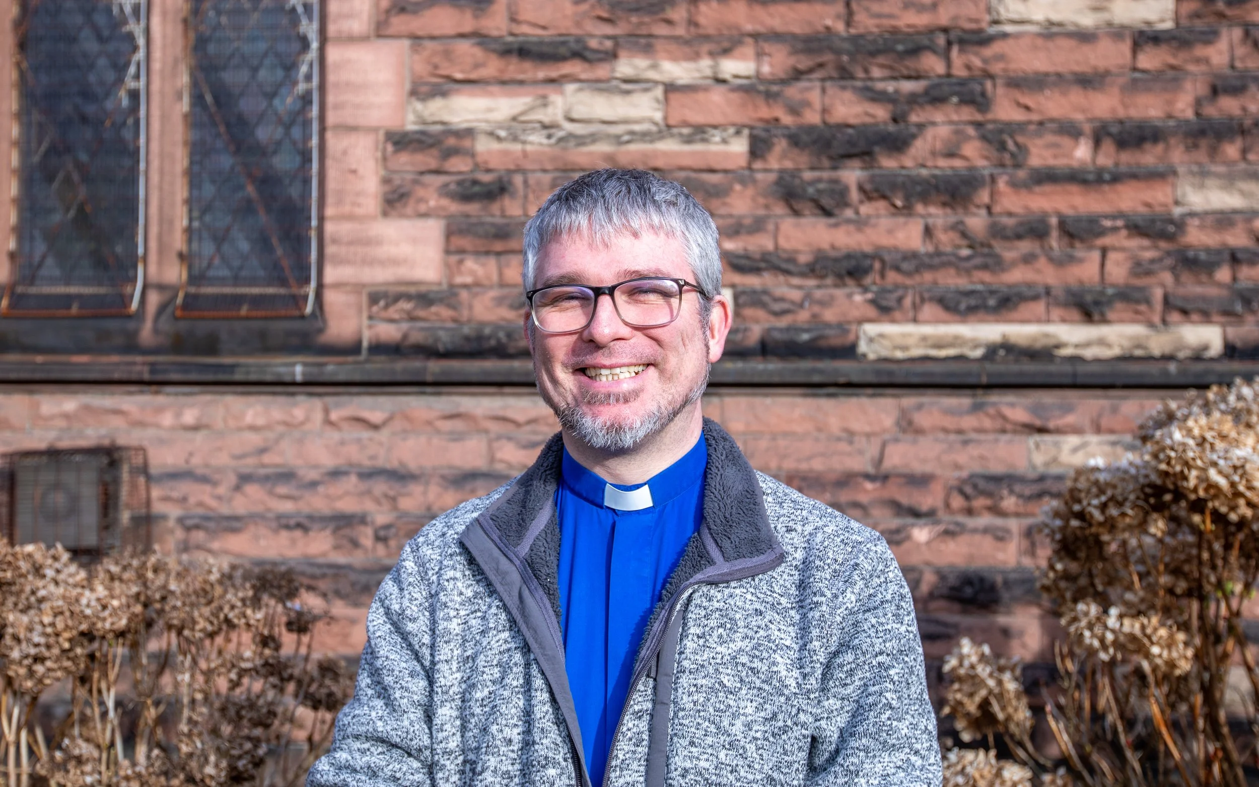 A smiling middle-aged man with glasses, gray hair, and a gray beard standing outdoors in front of a brick building with stained glass windows. He is wearing a blue clerical shirt with a white collar, a blue shirt, and a gray patterned zip-up jacket.