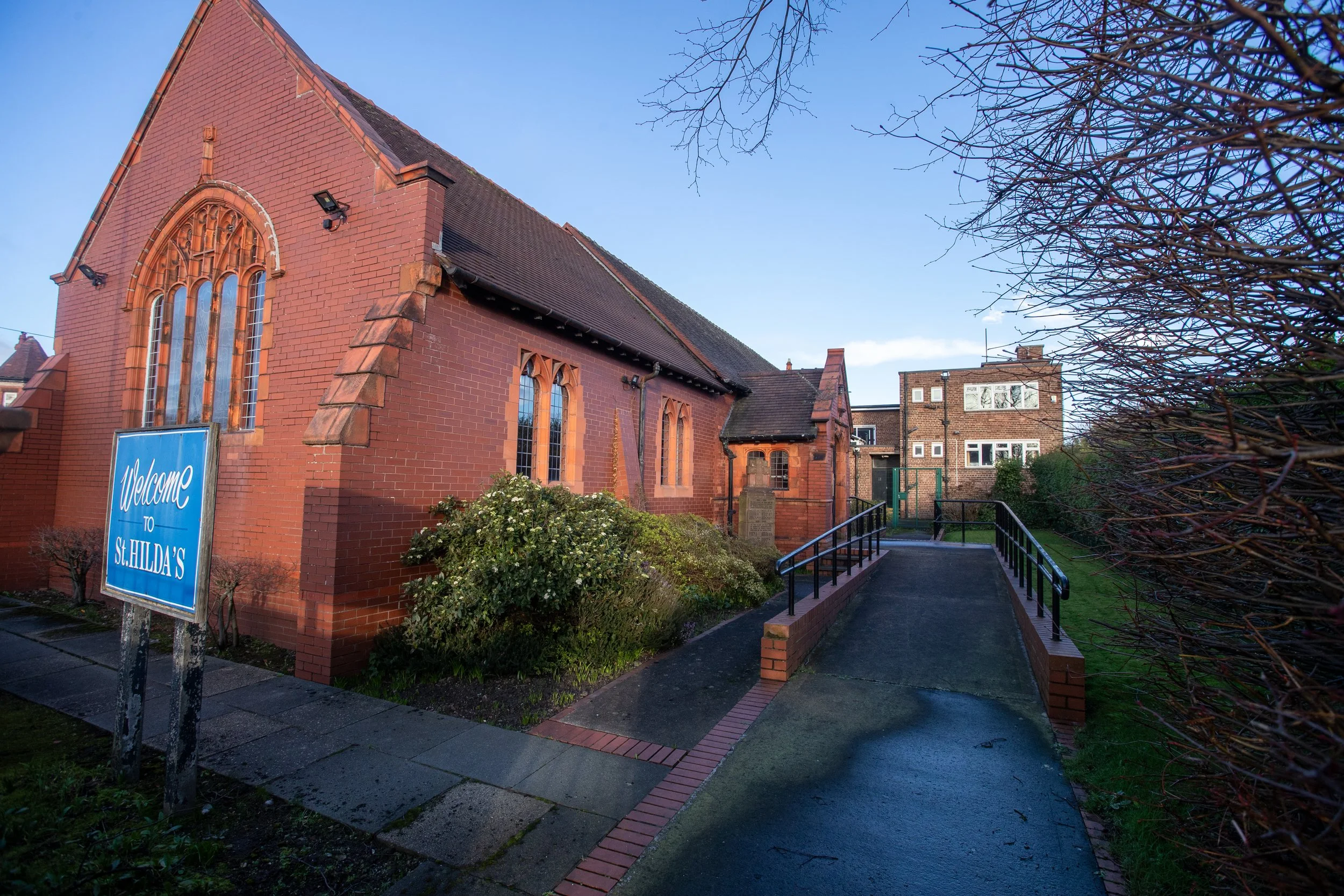 A red brick church with Gothic-style windows and a sign that reads 'Welcome to St. Hilda's'. The church is surrounded by bushes, trees, and a pathway with a black handrail leading to the entrance, under a clear blue sky.