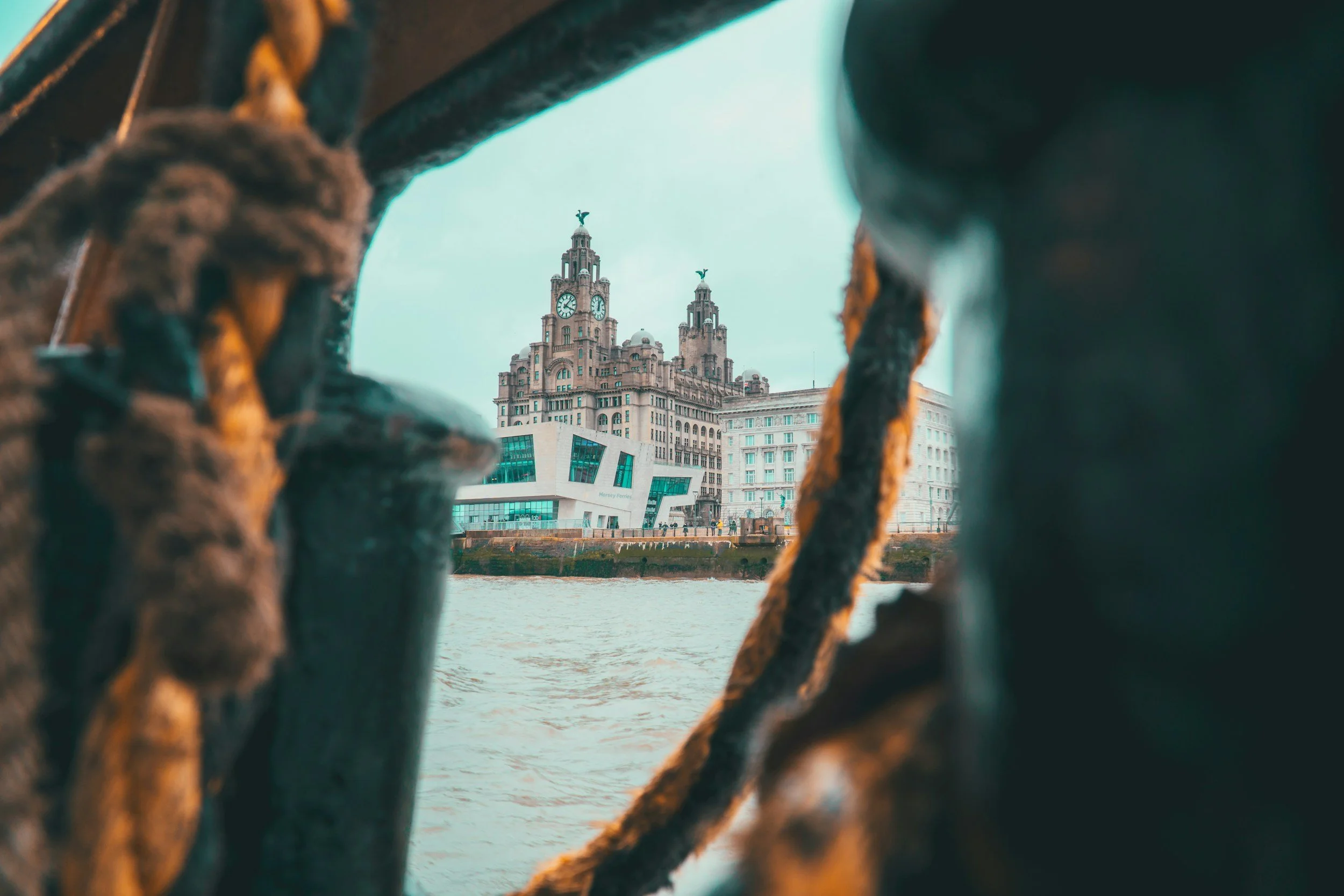 Looking through a rusted metal railing at the Royal Liver Building in Liverpool, England, with two clock towers topped with Liver birds, over the River Mersey.
