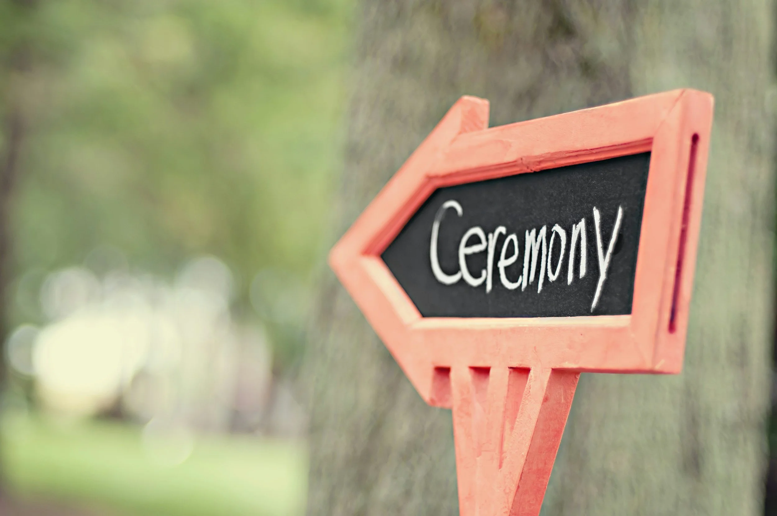 A small orange chalkboard sign with the word 'Ceremony' written on it in white chalk, mounted on a wooden signpost outside.
