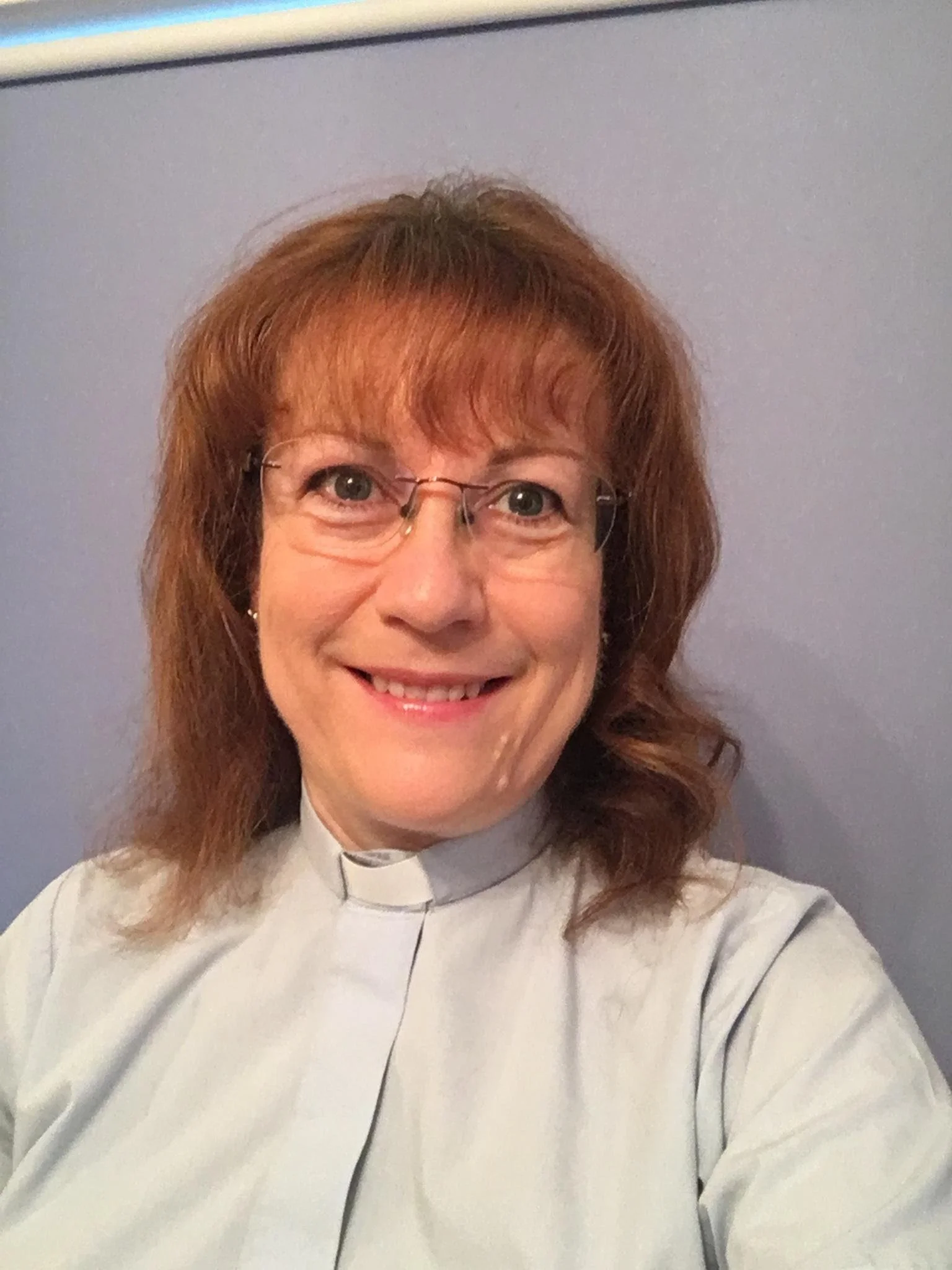 Smiling woman wearing glasses and a white religious clerical shirt with a clerical collar, standing in front of a gray wall.