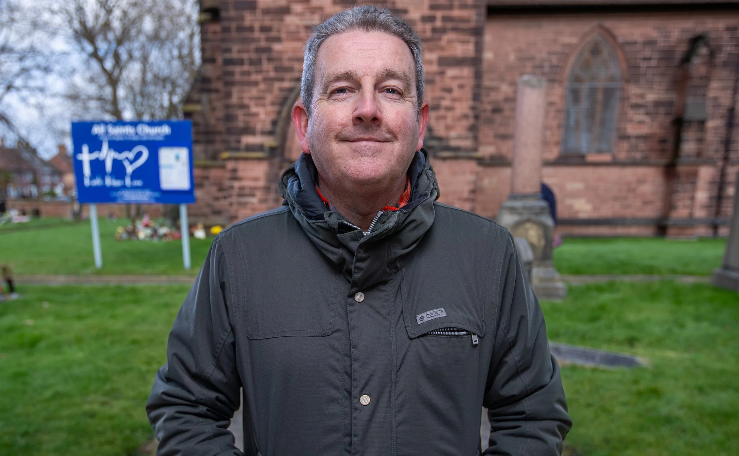 A man with short gray hair and blue eyes smiling outdoors in front of a brick church with arched windows. He is wearing a black jacket and standing on a grassy area. There are trees and a blue sign with white text and graphics in the background.
