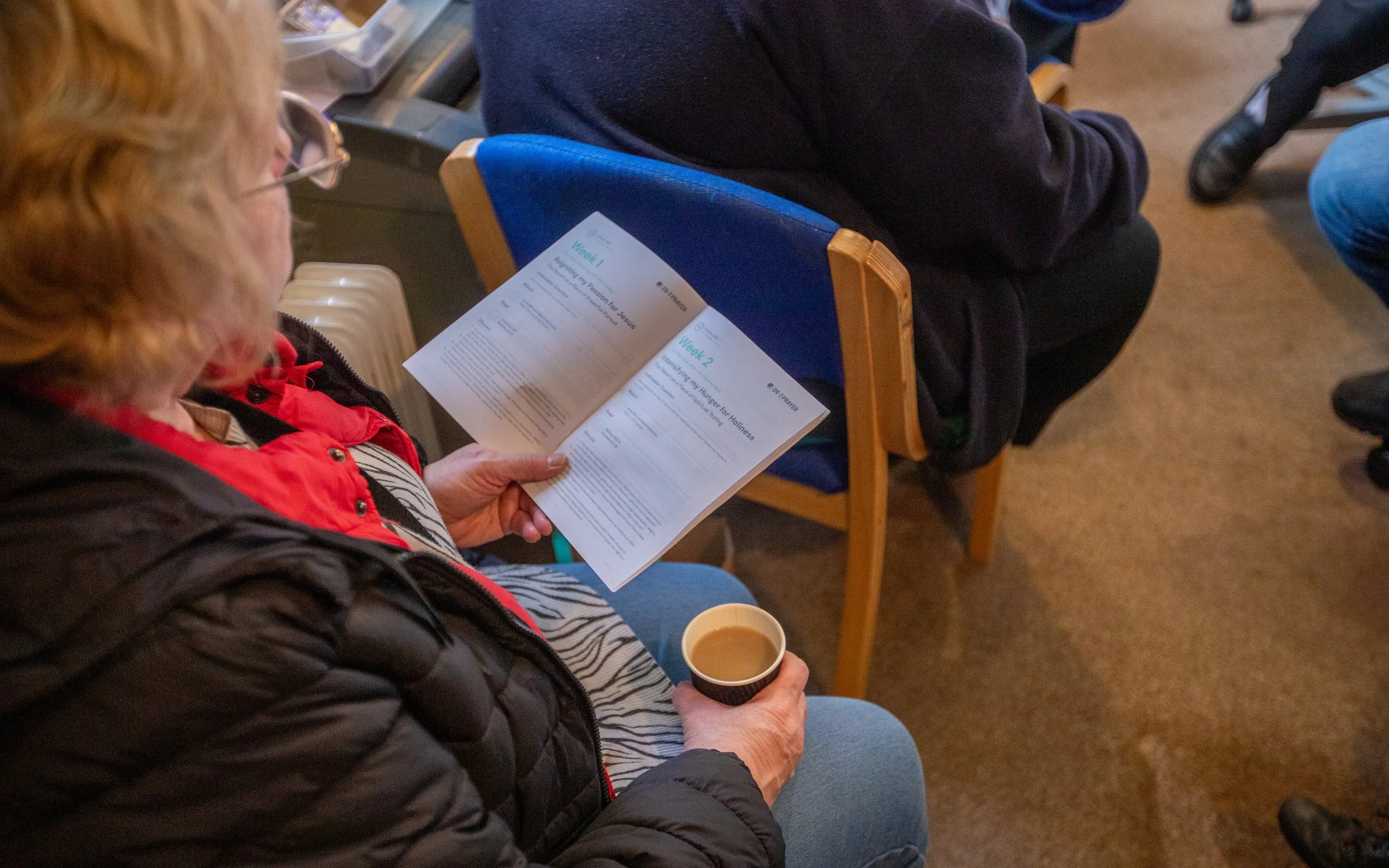 A woman in a black puffy jacket and red scarf is sitting on a blue-cushioned wooden chair, reading a booklet titled 'Week 1' and 'Week 2' about spiritual topics. She holds a small paper cup of coffee in her right hand, and there are other people sitting nearby in this indoor setting.