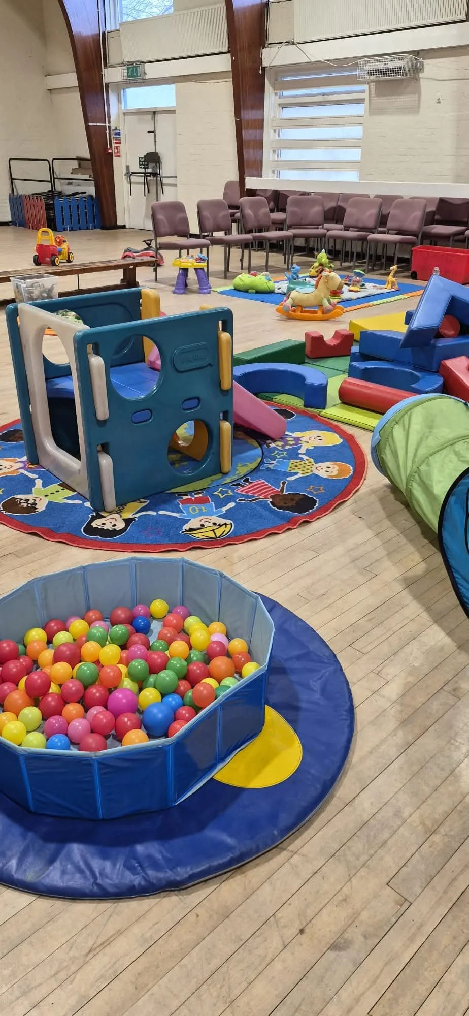 Indoor children's play area with colorful plastic ball pit, slide, and various toys. Also contains chairs along the wall and a blue padded mat with a circle of brightly colored plastic balls.