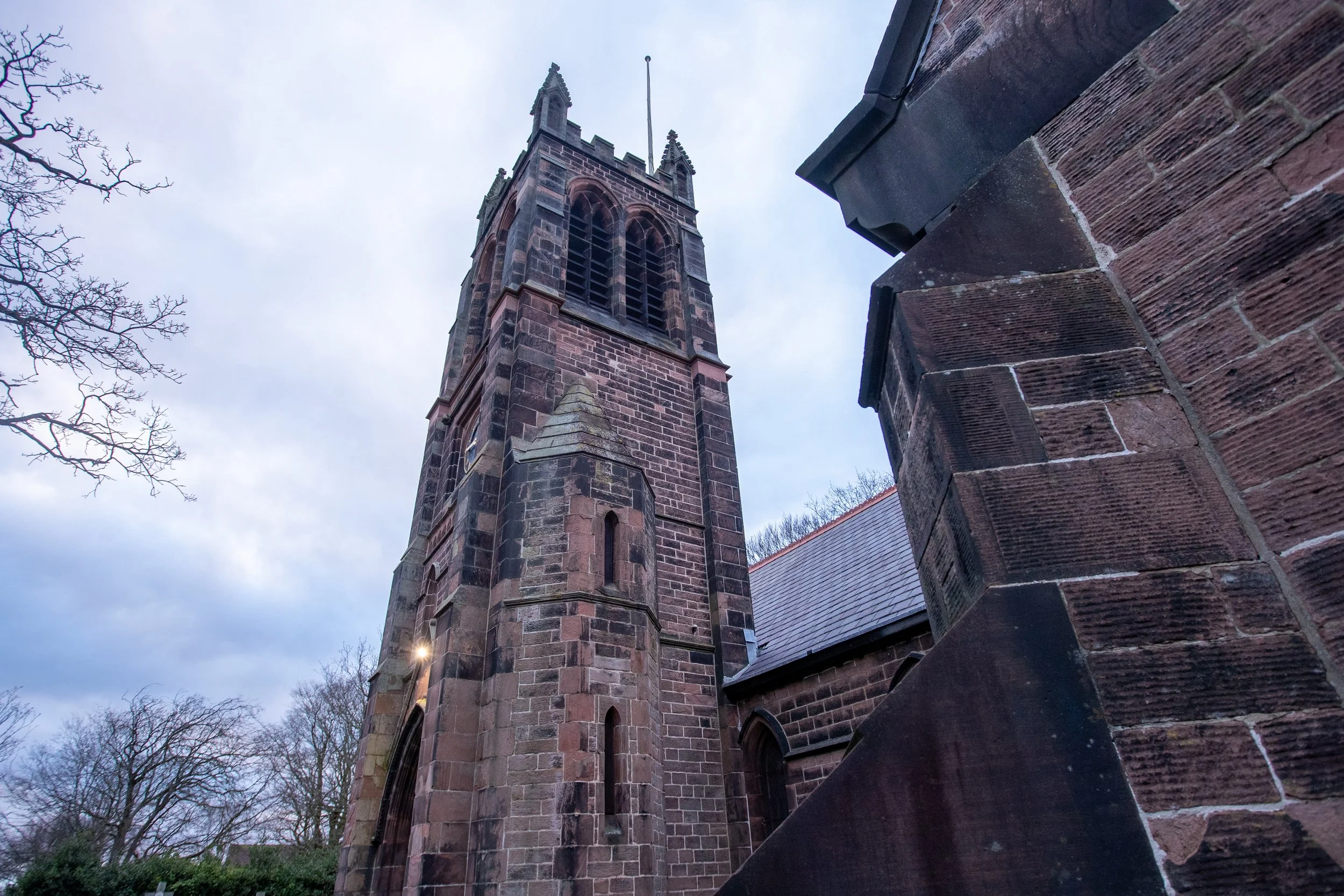 A tall brick church steeple viewed from the ground, with an overcast sky and leafless trees in the background.