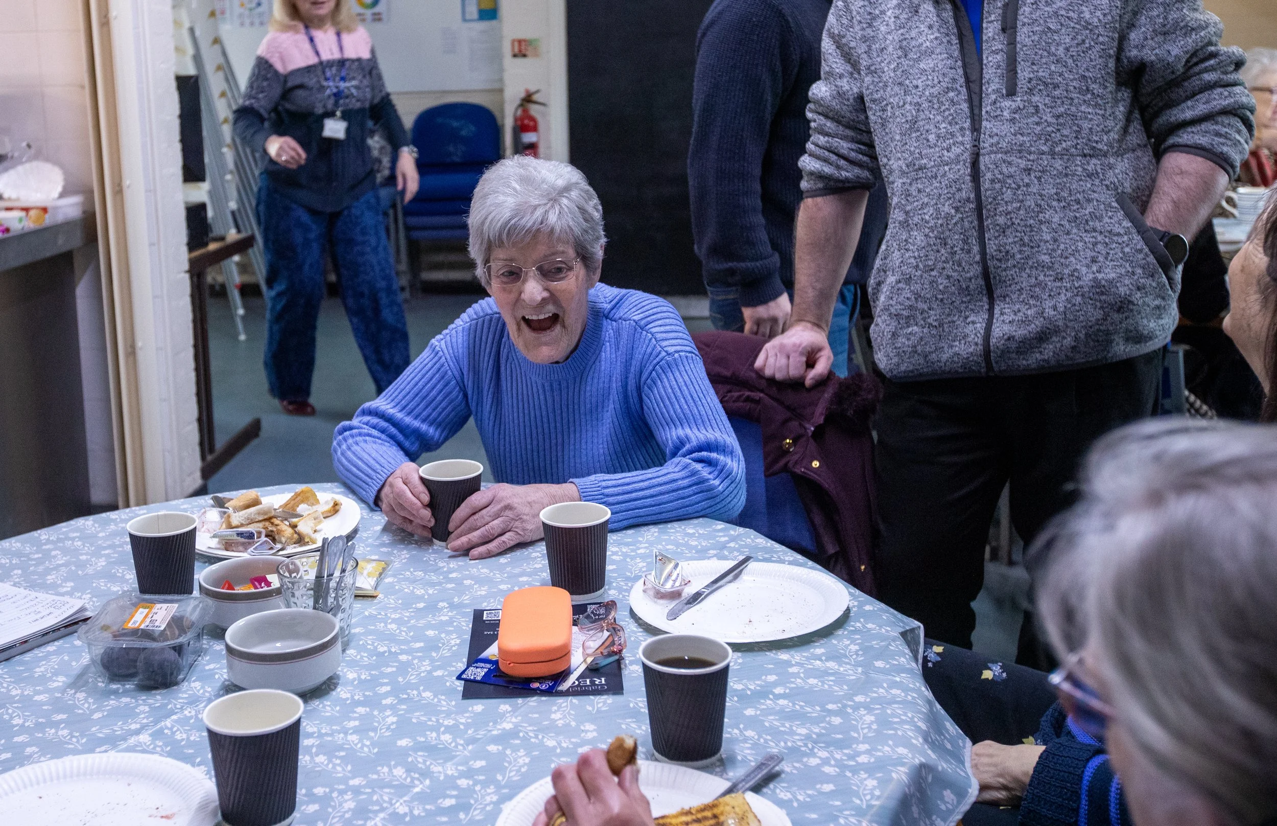 An elderly woman with glasses and short gray hair sitting at a table, smiling with her mouth open, holding a black cup. The table is decorated with a blue and white patterned tablecloth, and has plates with food, cups, and utensils. Several other people are standing or sitting nearby in a room with chairs, decorations, and a fire extinguisher.