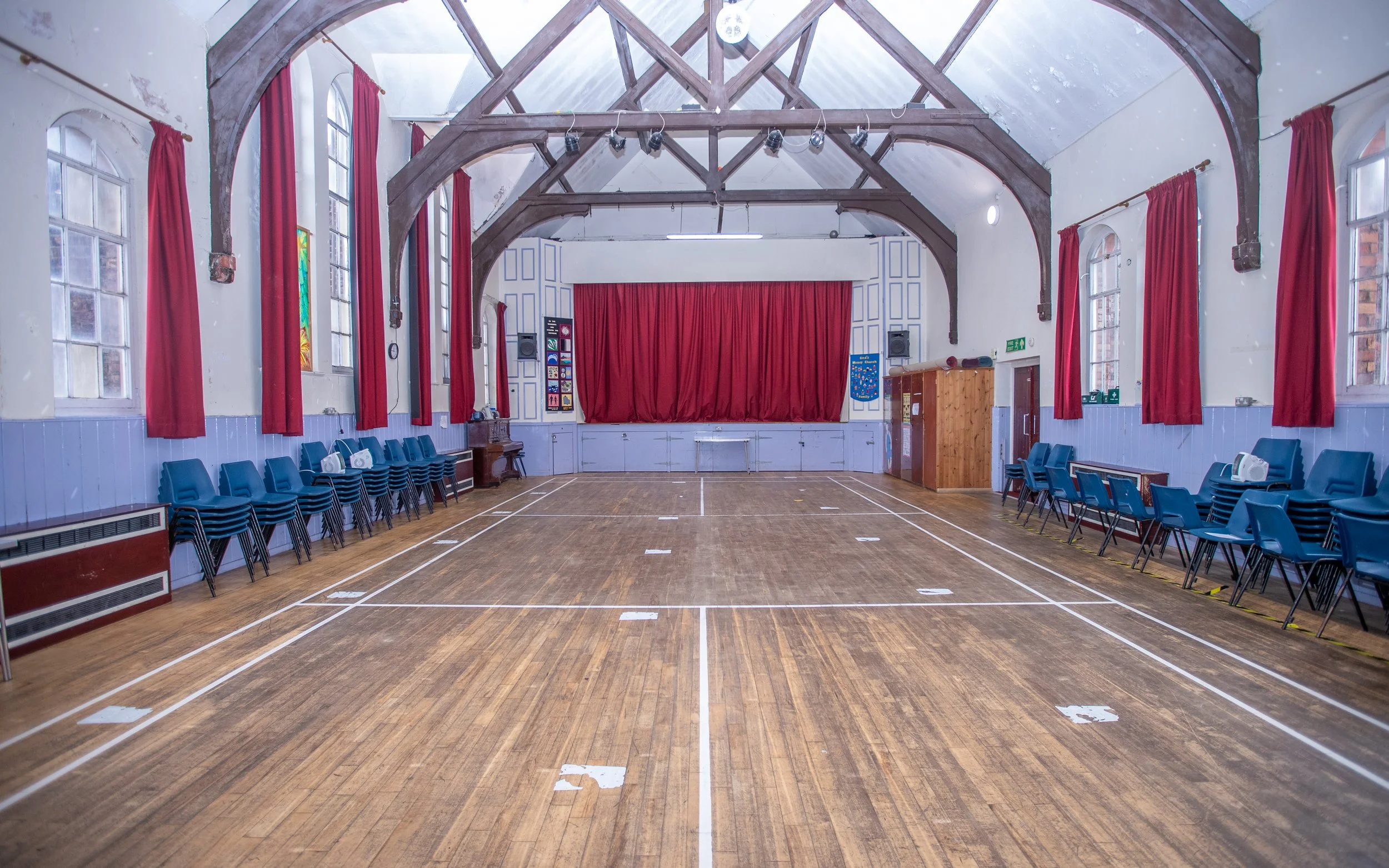 An empty indoor gymnasium with a wooden floor, red curtains on large windows, a red stage curtain, blue chairs arranged along the walls, and wooden beams on the ceiling.