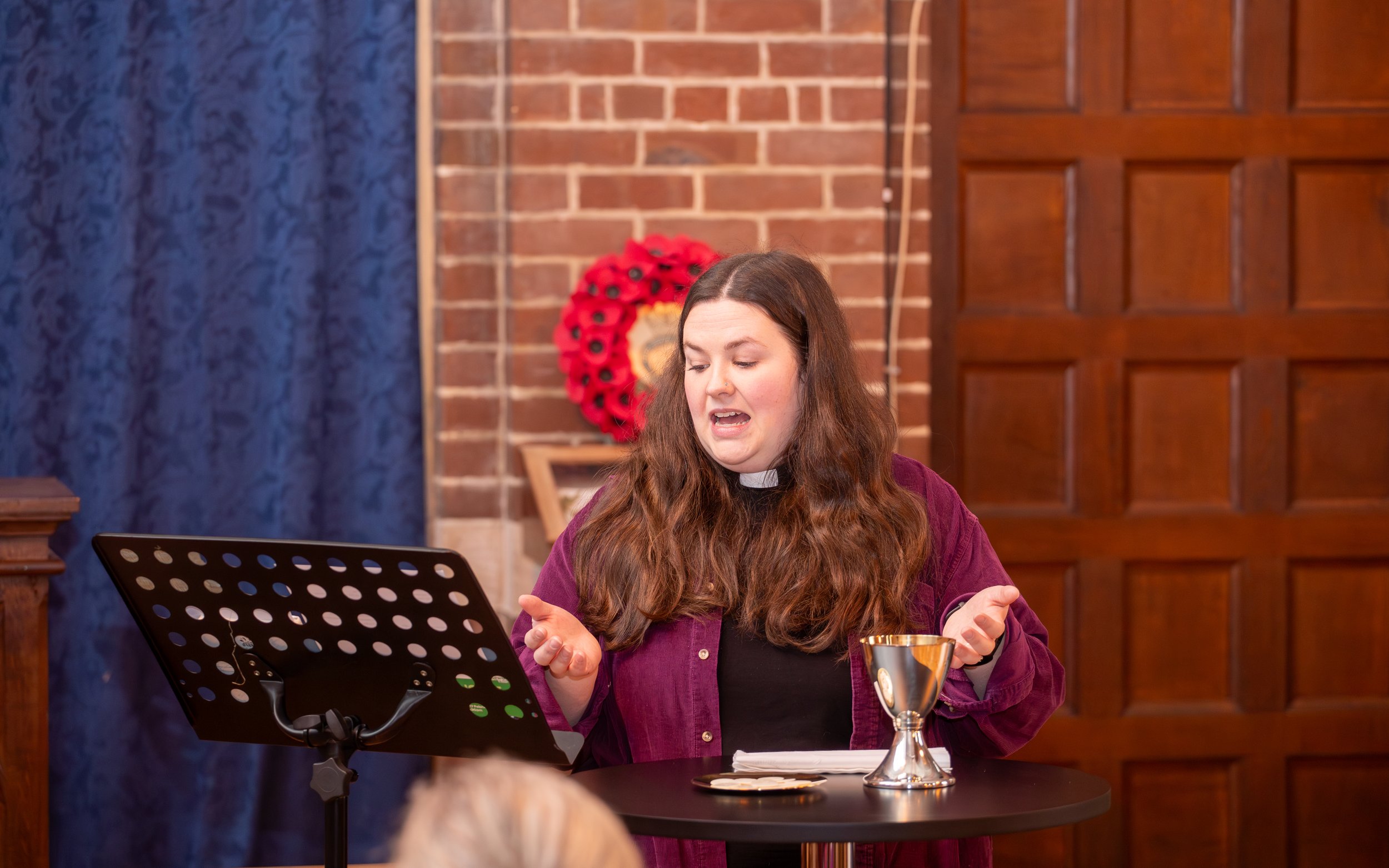 A member of clergy with long brown hair leading communion. Behind her are brick and wood-paneled walls, decorated with a red poppy wreath.