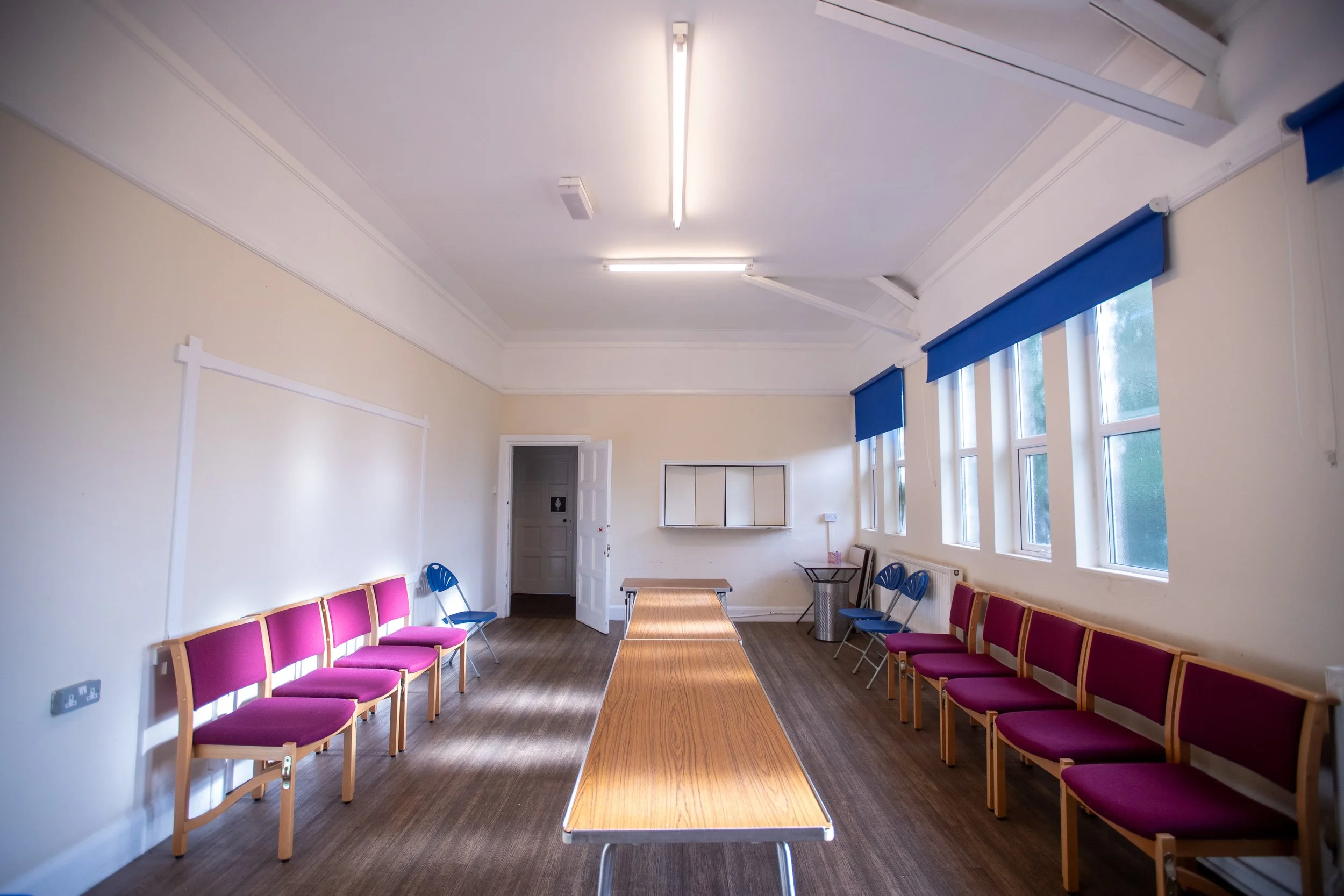 A room with beige walls, a wooden table in the center, pink and blue chairs along the walls, windows with blue blinds, and ceiling lights.