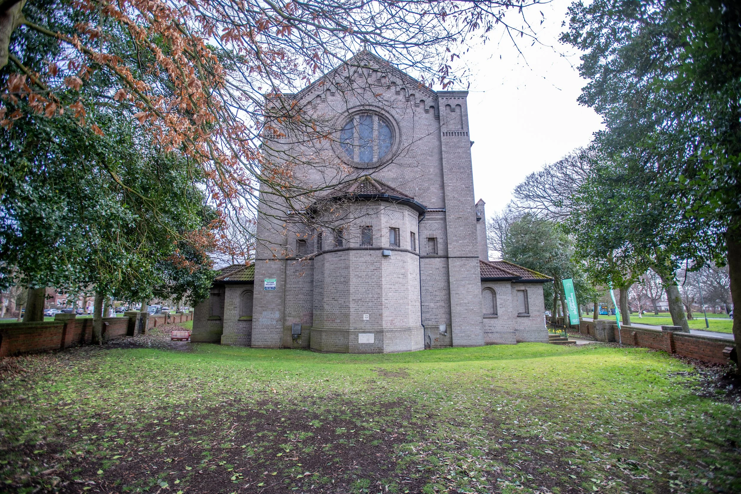 A brick church with a large circular stained glass window, surrounded by trees with bare branches and some green leaves, in a park setting with grass and pathways.