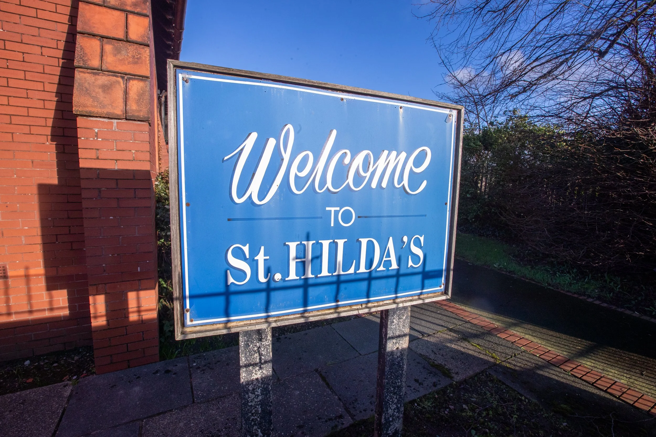 Blue welcome sign with white text that reads 'Welcome to St. Hilda's,' mounted on concrete posts outside near a red brick building and trees with some branches.