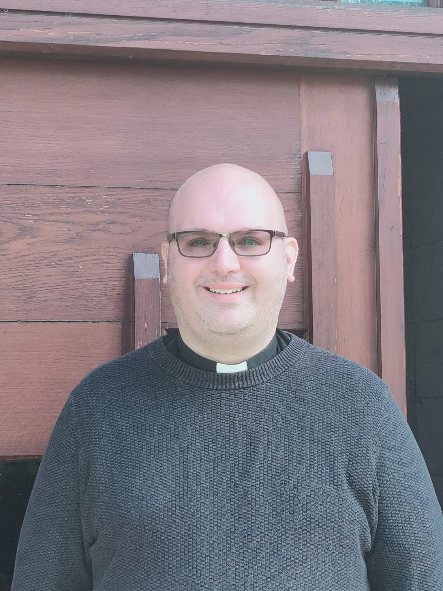 A man with glasses, smiling, wearing a black sweater with a clerical collar, standing in front of a wooden wall.