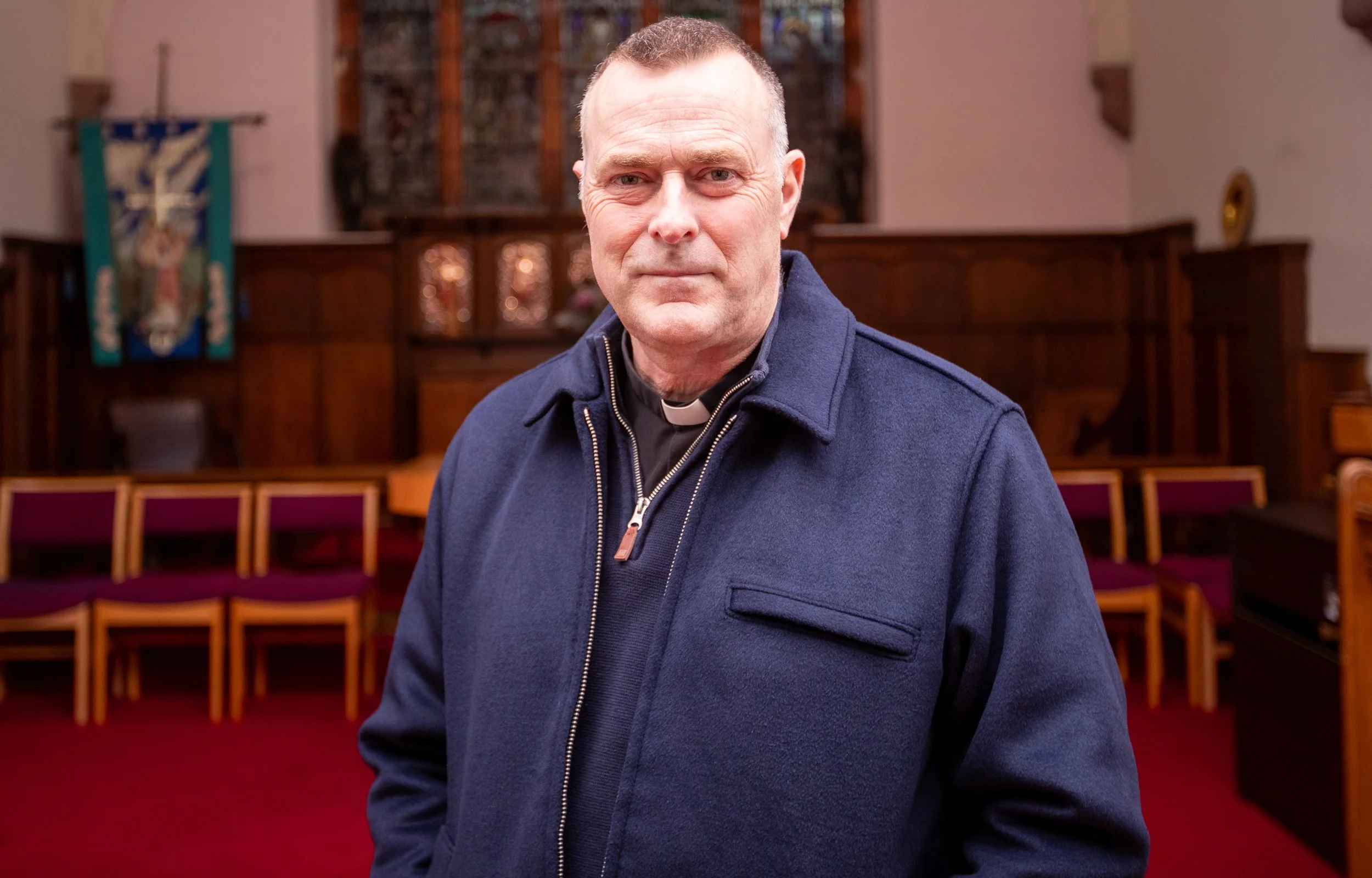 A man standing inside a church, wearing a dark blue jacket and smiling slightly, with rows of red chairs and wooden church interior in the background.