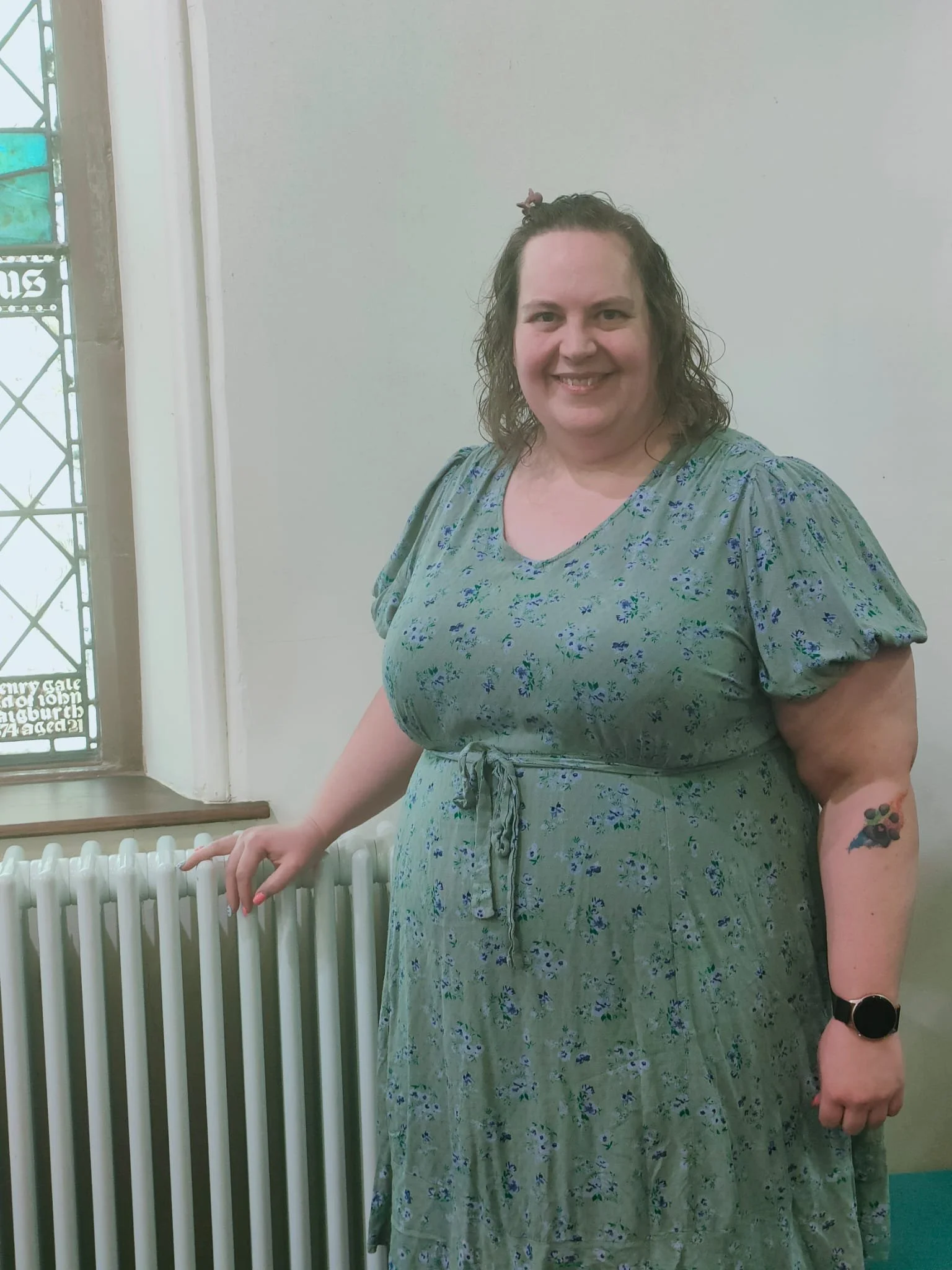 A woman standing next to a radiator on a light-colored wall. She is smiling, wearing a green floral dress, a black watch, and has a colorful tattoo on her left arm. Her hair is curly and tied up with a pink scrunchie.