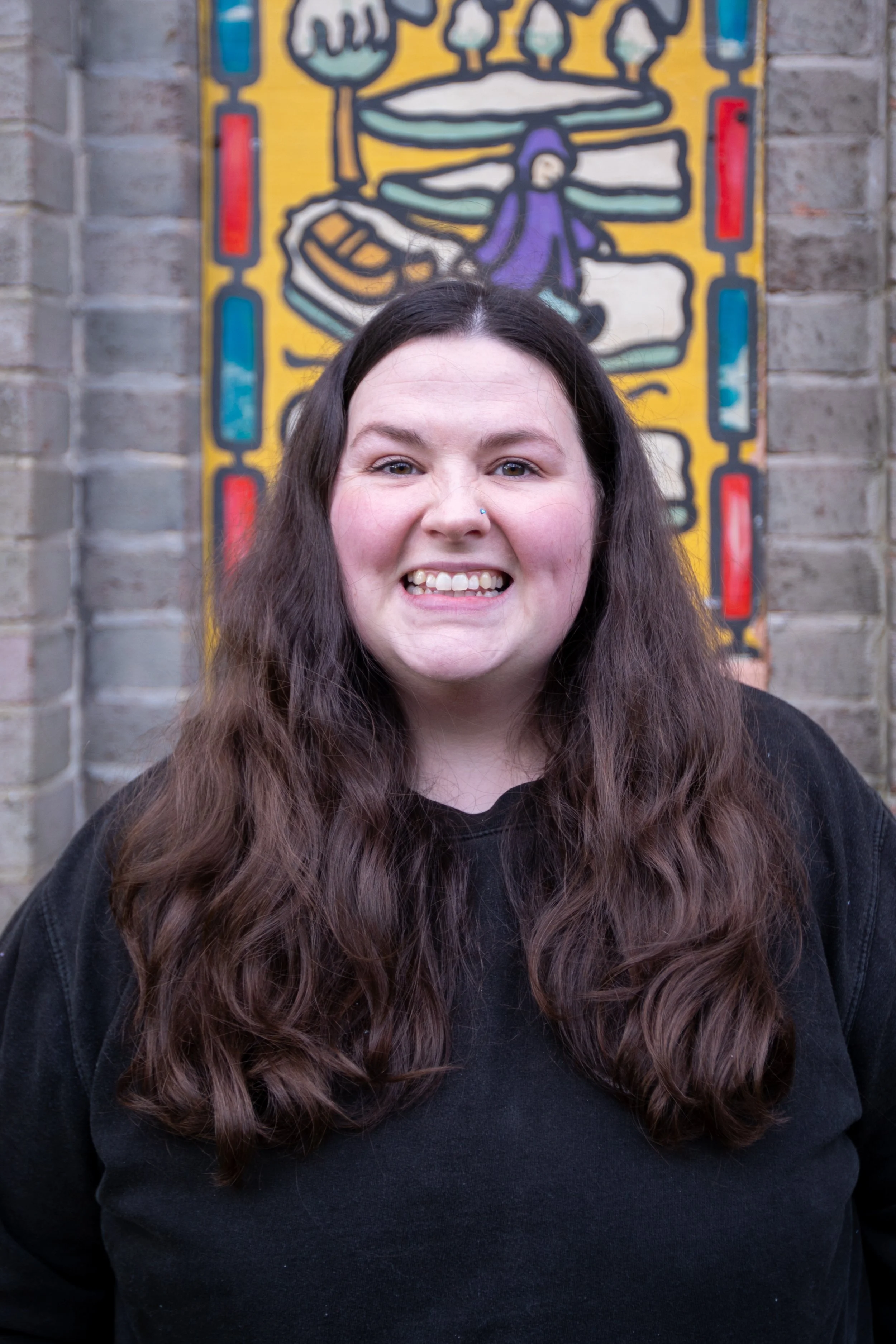 A woman with long, wavy brown hair and a nose piercing smiling in front of a colorful mural on a brick wall.