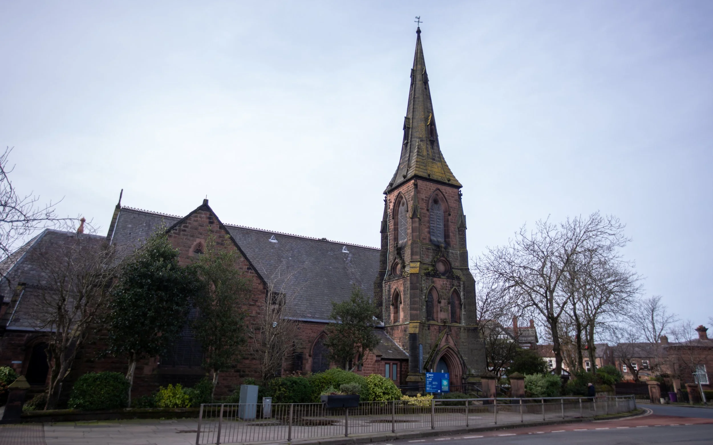 A historic church made of reddish-brown stone with a tall, pointed steeple topped with a weather vane. The church is surrounded by leafless trees and a small garden, with a street and sidewalk in the foreground.