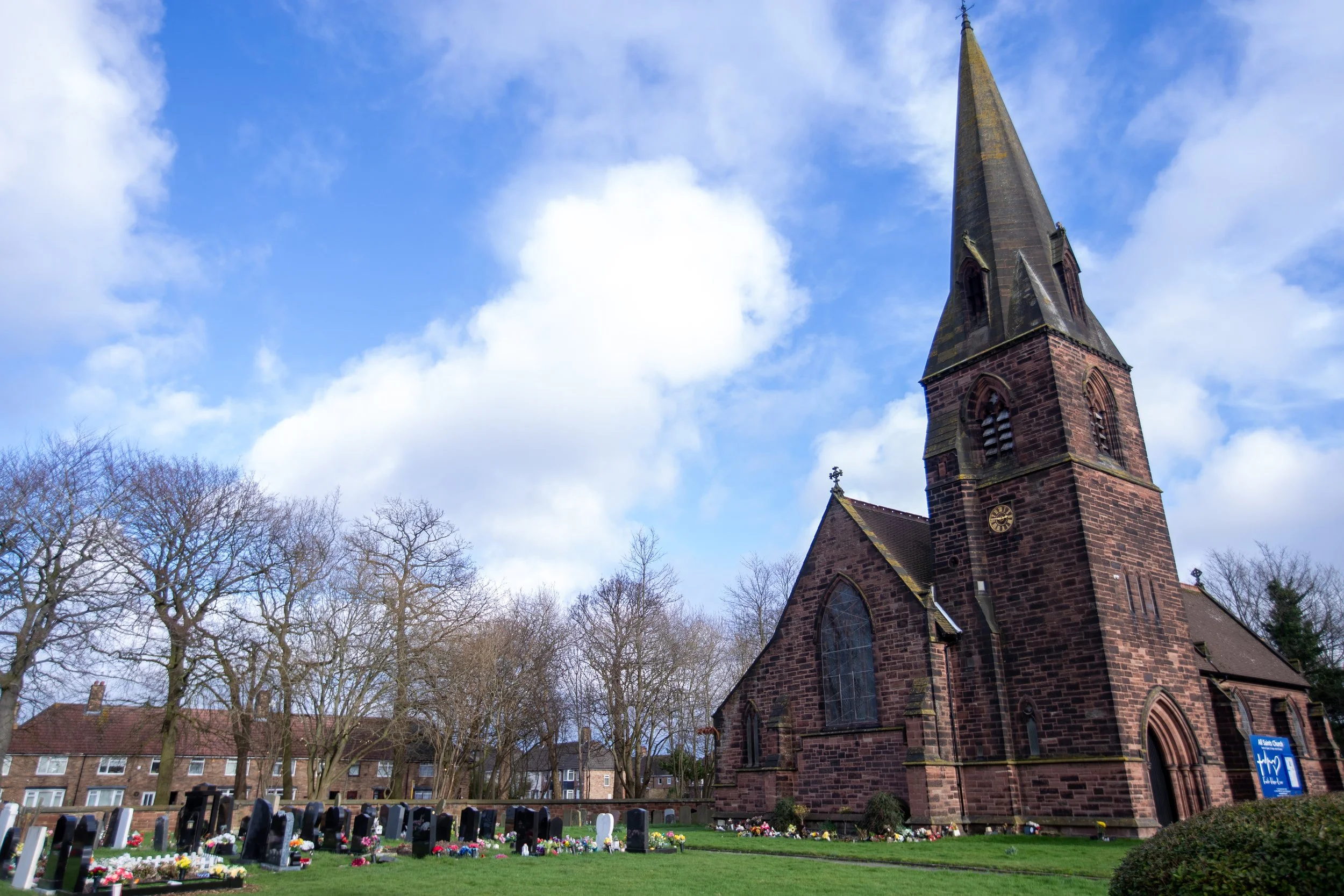 A stone church with a tall steeple and a clock, surrounded by a cemetery with graves and trees, under a partly cloudy sky.