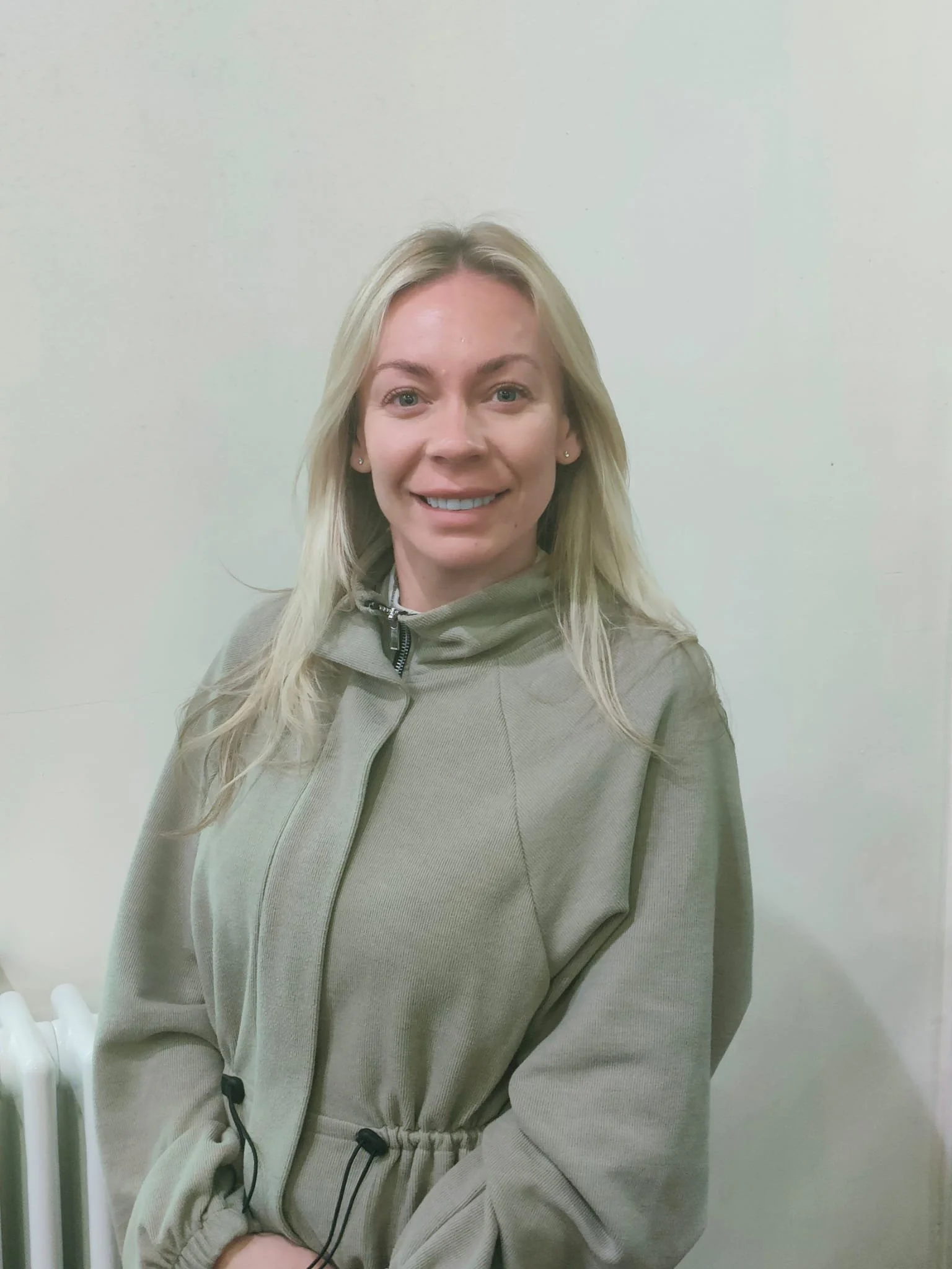A woman with long blonde hair, wearing a light gray hoodie, smiling and sitting against a plain light-colored wall.