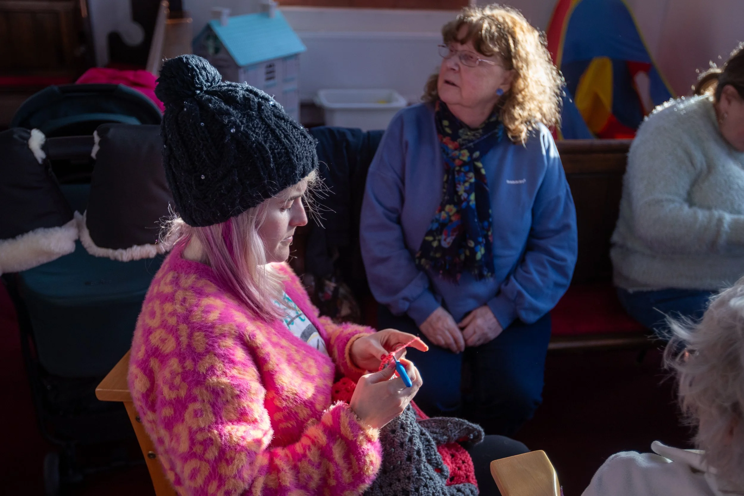 A young girl with pink hair and a black knit hat sandbar a pink and orange fuzzy sweater, sitting and knitting while a woman with curly hair wearing glasses and a blue jacket looks on in a colorful indoor play area with a tent and toys in the background.