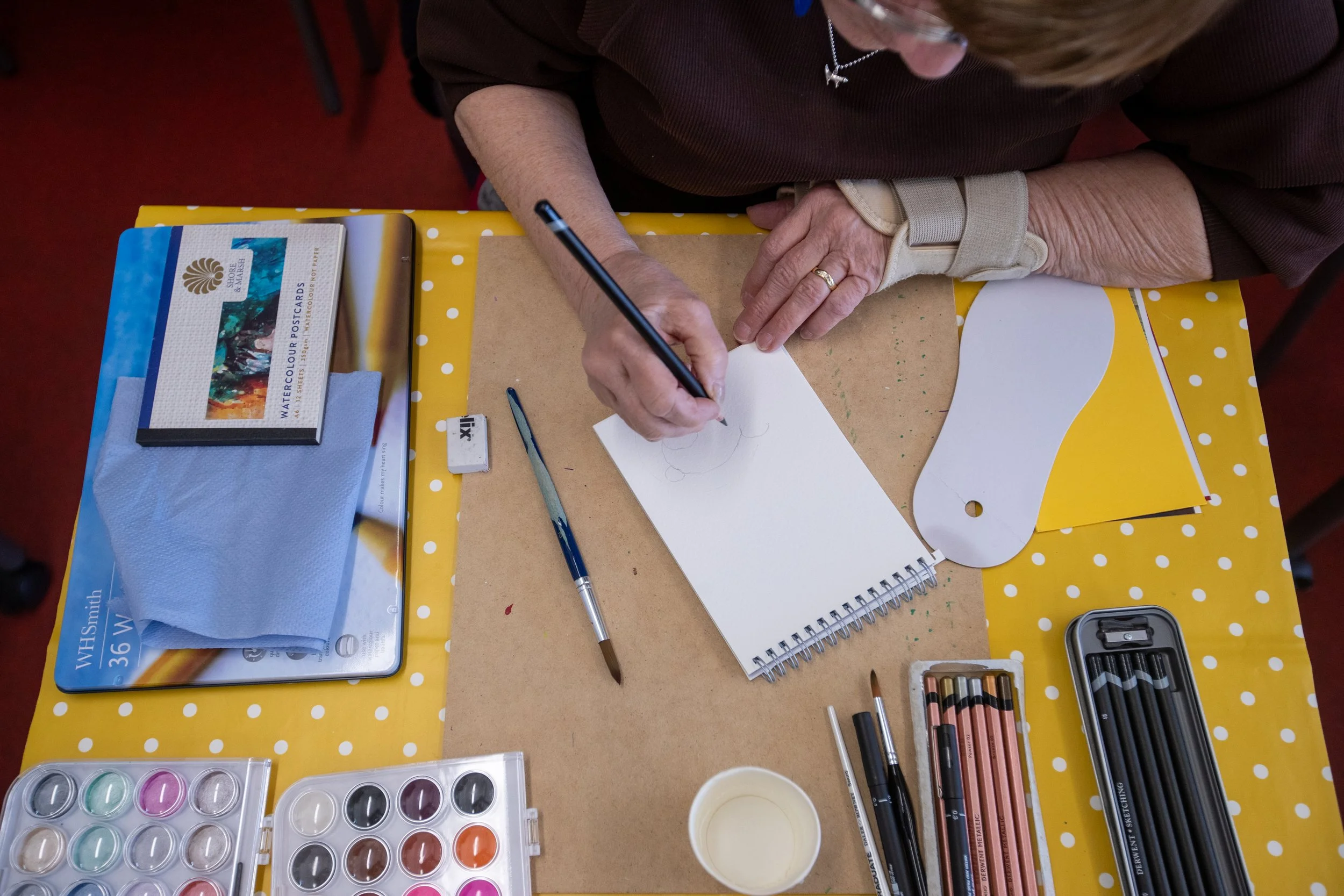 Overhead view of a person drawing on a notepad with colored pencils and markers on a yellow polka dot table. Art supplies including watercolor paint set, brushes, eraser, and paper sheets are also visible.