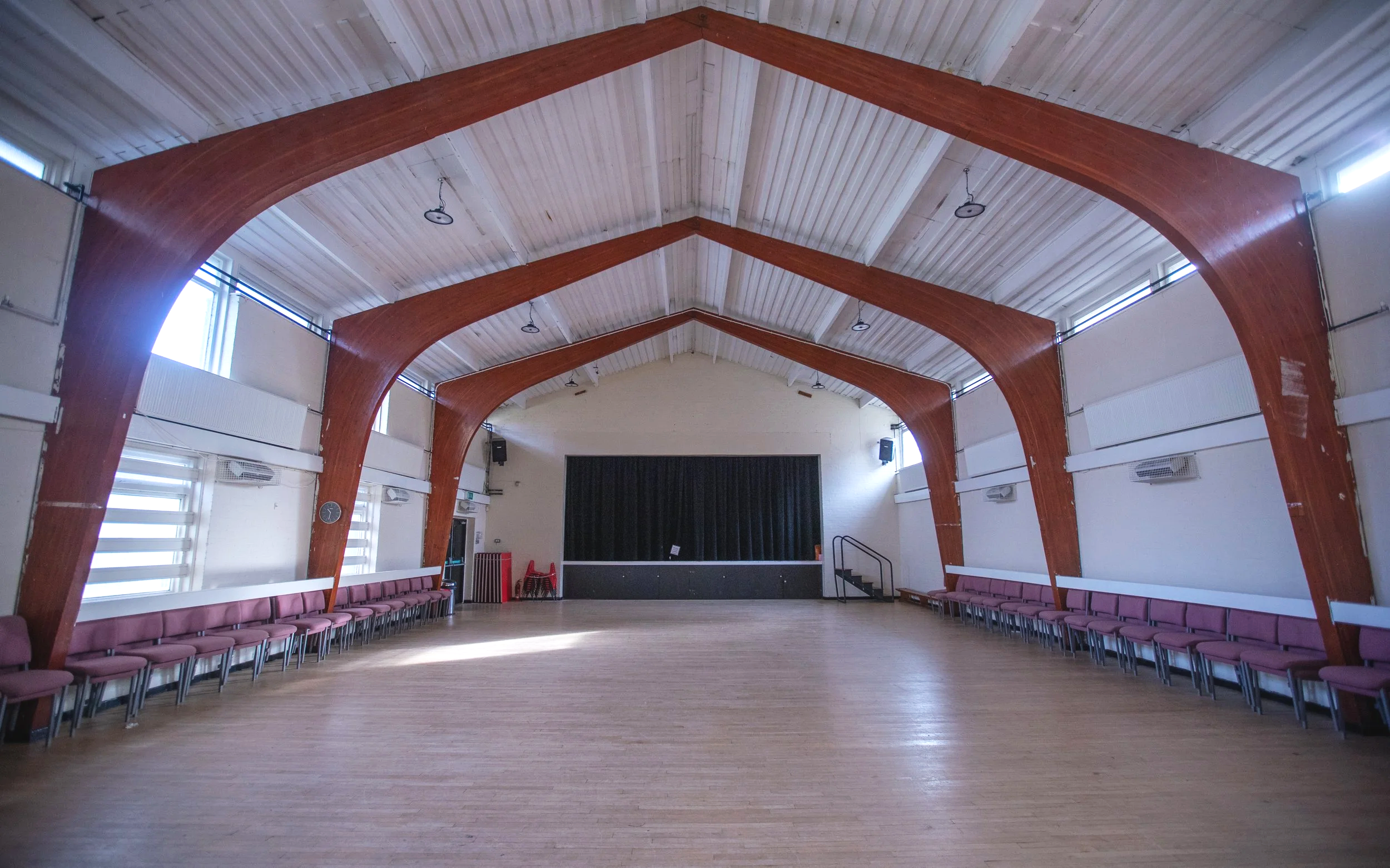 An empty indoor event hall or auditorium with a wooden vaulted ceiling, rows of maroon chairs along the sides, and a stage with black curtains at the far end.