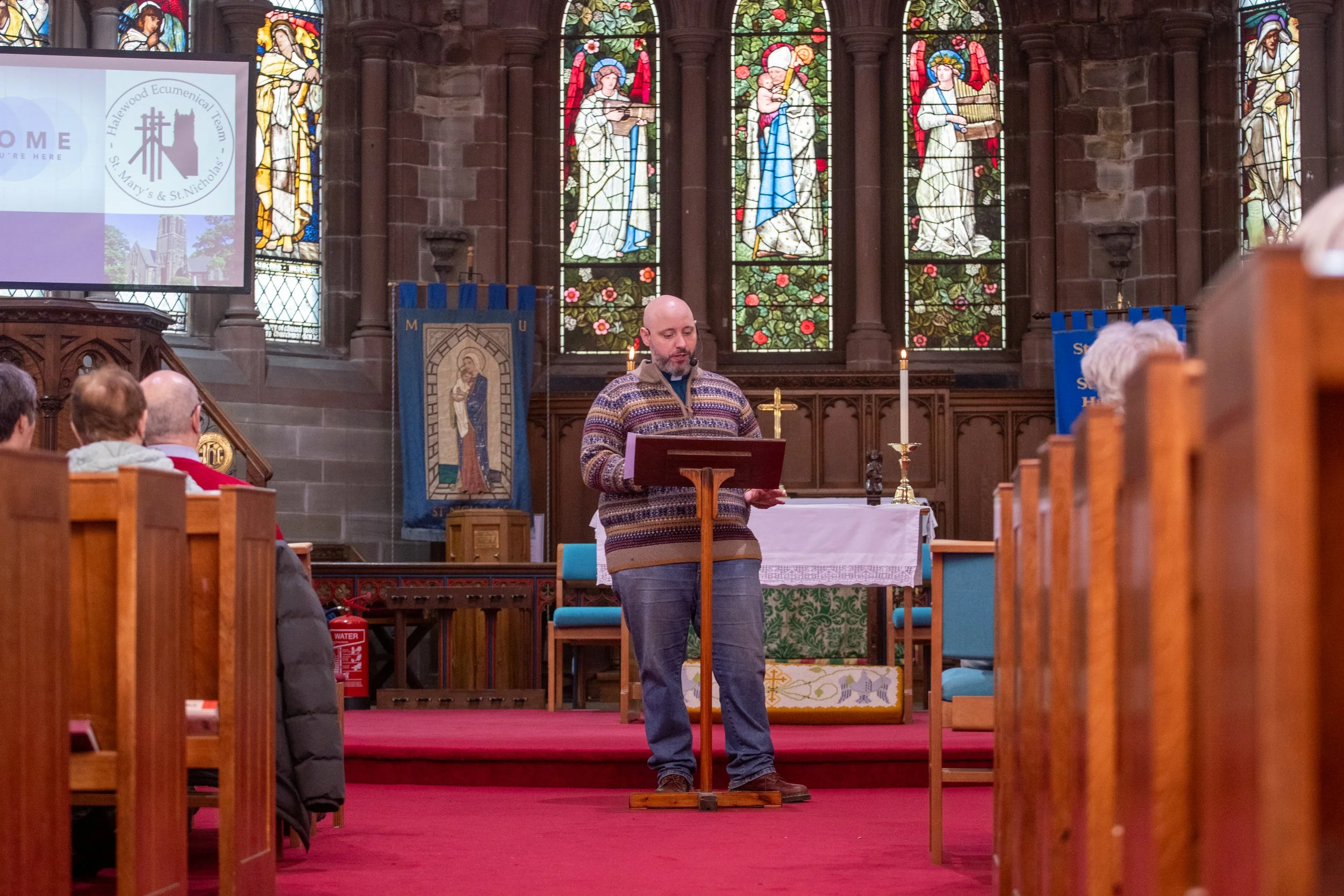 A man standing at a pulpit inside a church with stained glass windows in the background. Several people are seated in the pews, and there is a cross and candles on the altar. The church has red carpet and wooden decor.