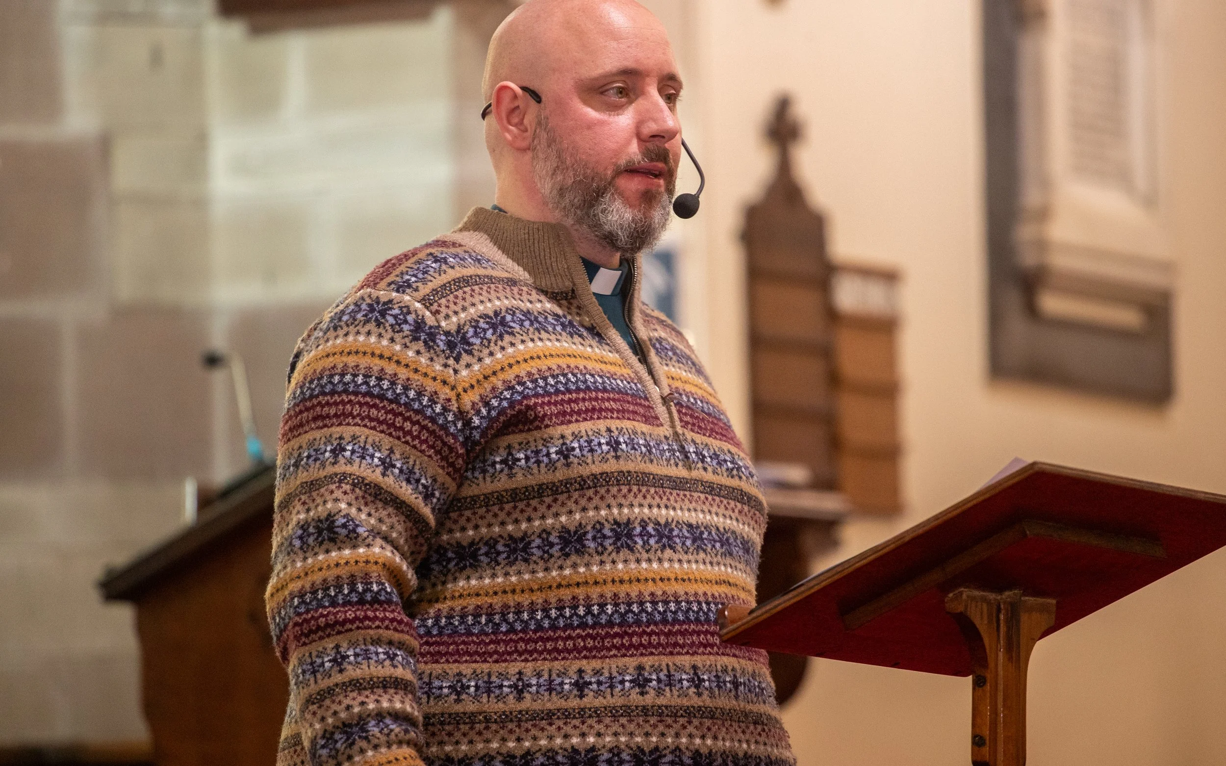 A member of clergy with a beard and wearing a colourful, patterned sweater, standing in a church, reading from a lectern.