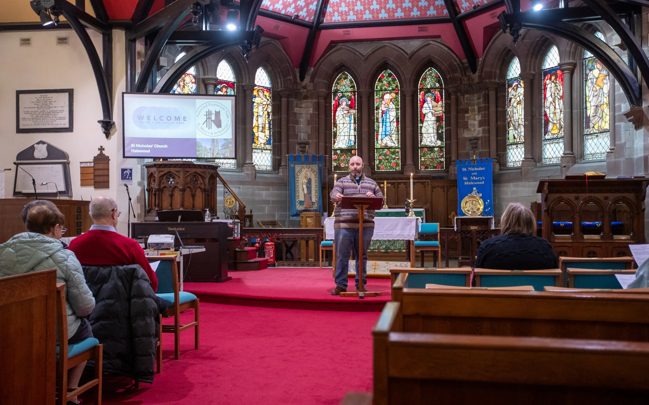 Interior of a church with stained glass windows depicting religious figures. A man is standing at a pulpit, addressing the congregation. Several people are sitting in pews, listening, with some using devices or reading. There is a large screen displaying a welcome message for St. Nicholas' Church in Halewood.