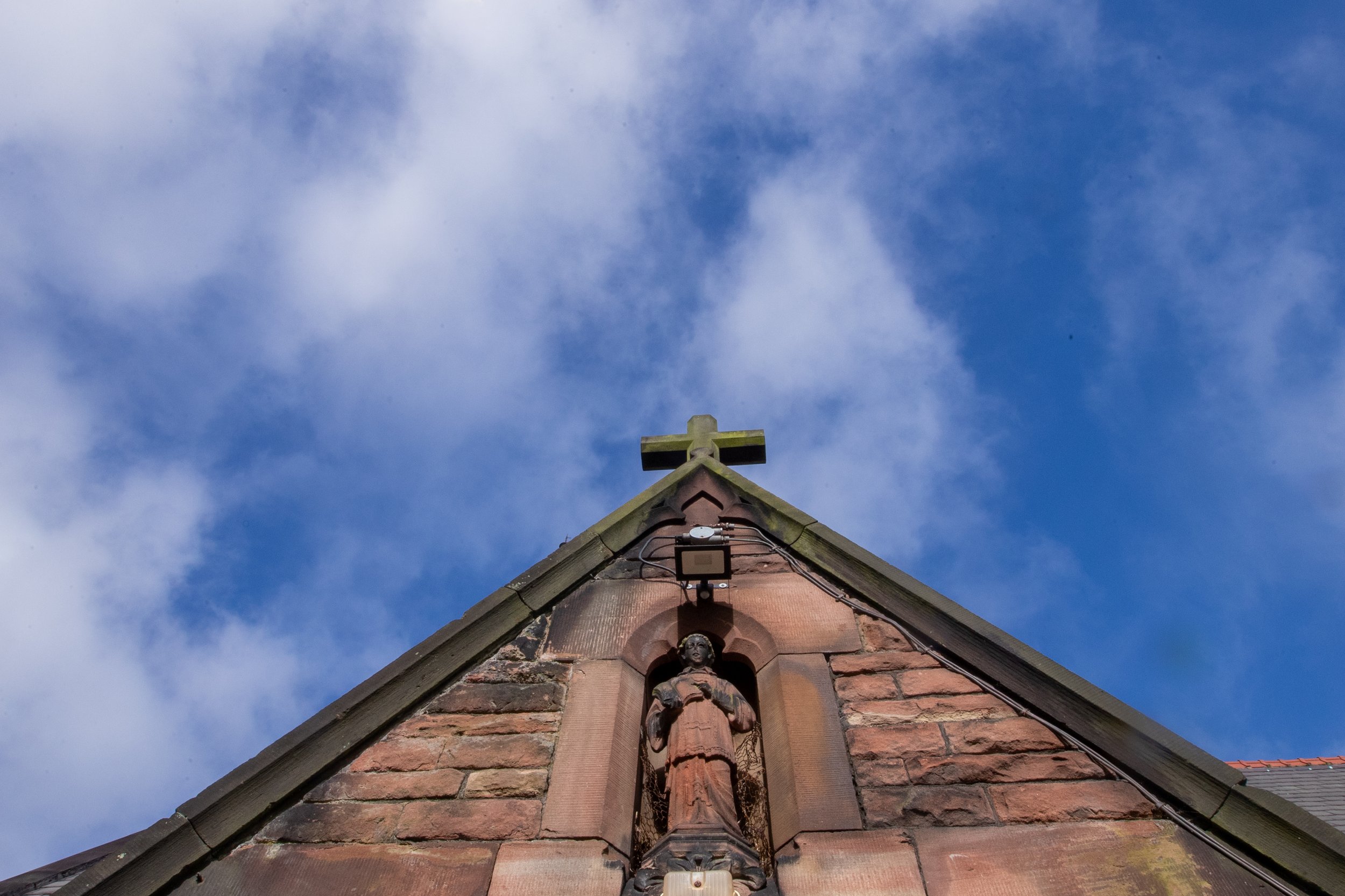 Looking up at the roof of a church or historic building made of red bricks, with decorative statues and a cross at the top, against a partly cloudy blue sky.