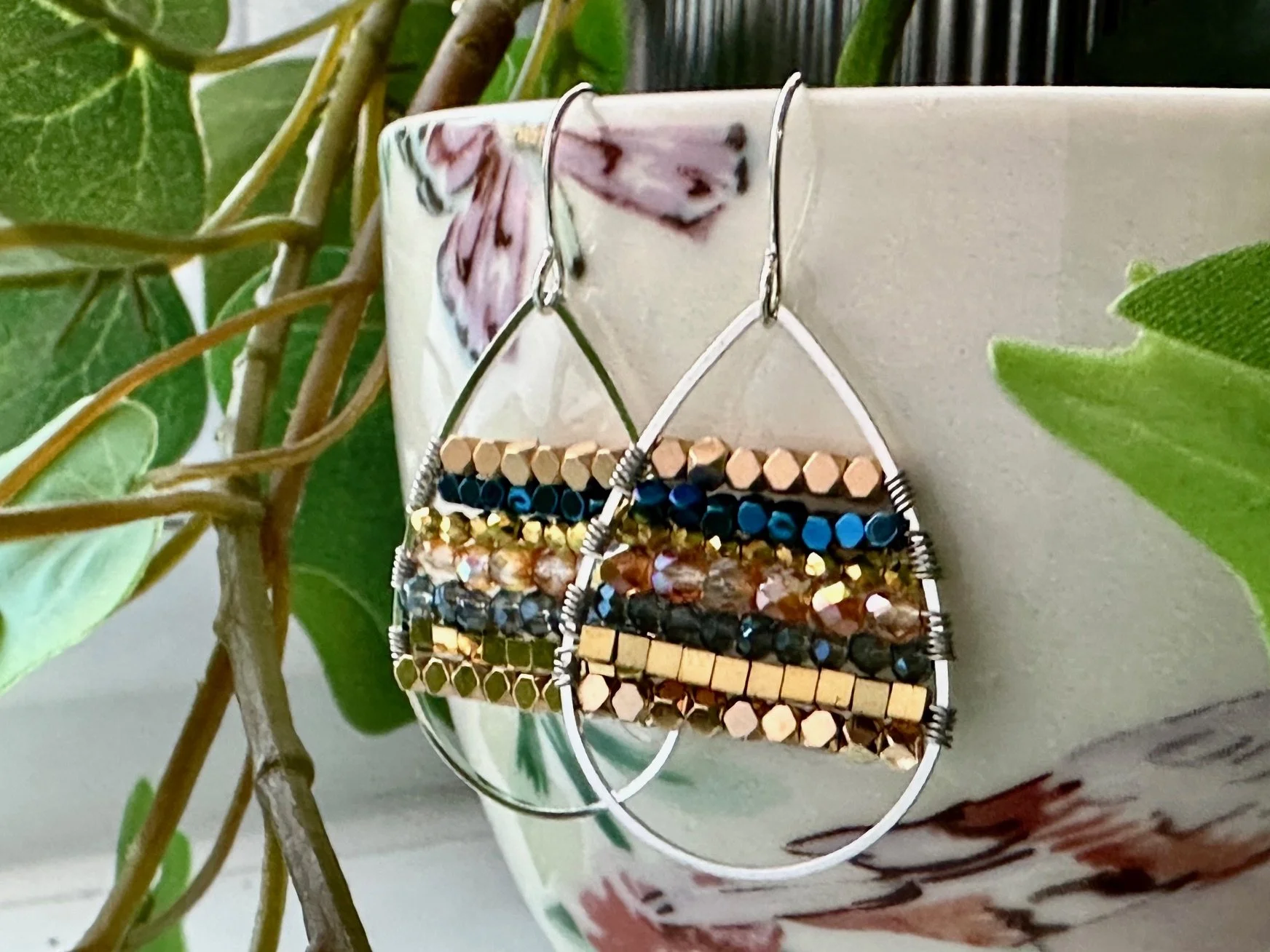 Colorful beaded earrings hanging on the edge of a floral-patterned ceramic pot with green leaves and stems nearby.