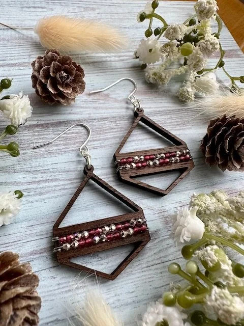 Wooden geometric earrings with pink and silver beads surrounded by pinecones, white and green flowers, and dried plants on a white wooden surface.
