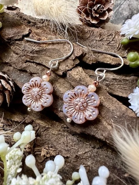 Pink flower-shaped earrings with pearl and bead accents, hanging on a wooden background with white and brown flowers around.
