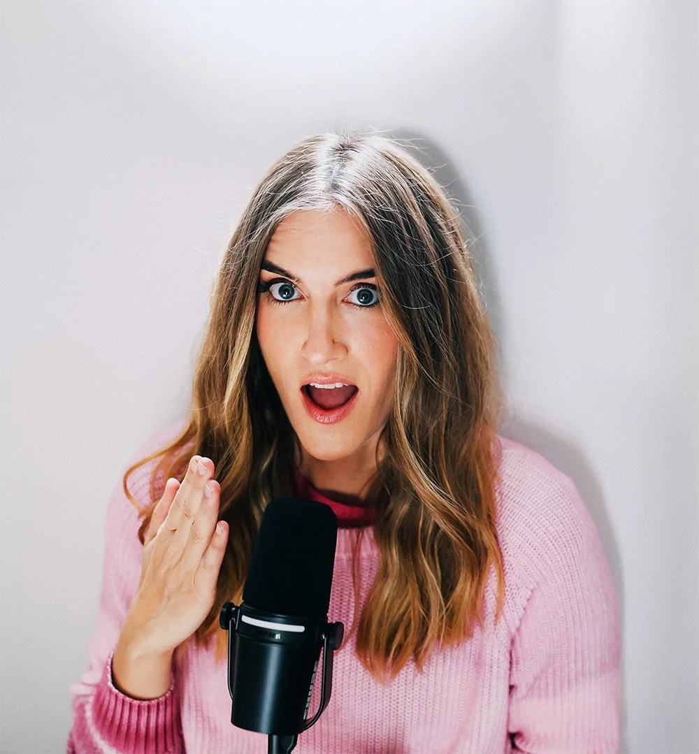 A woman with long, wavy brown hair and blue eyes gestures with her hand while singing or speaking into a black microphone, wearing a pink sweater, against a plain white background.